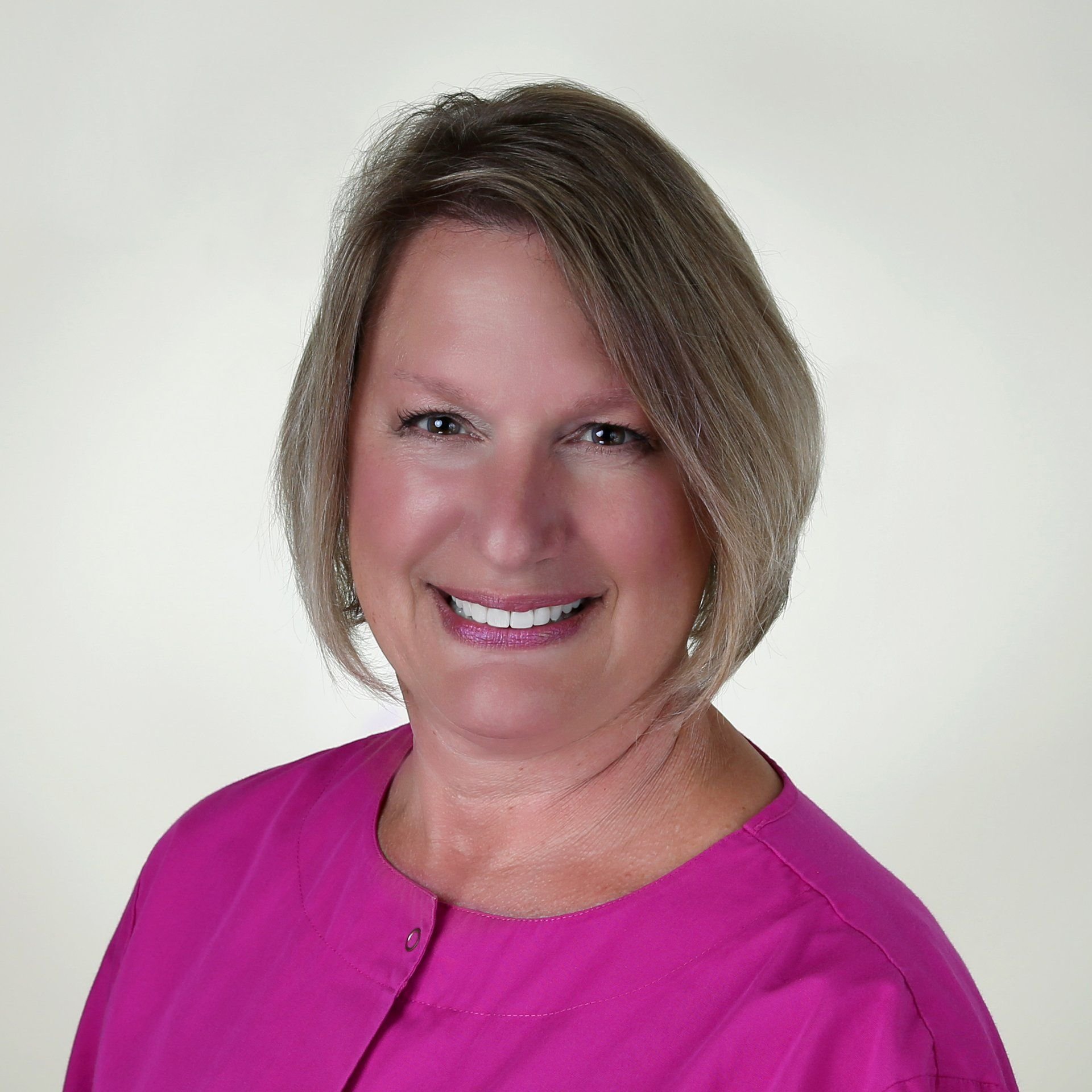 Portrait of a smiling woman with short blonde hair, wearing a bright pink top, against a plain, light-colored background.