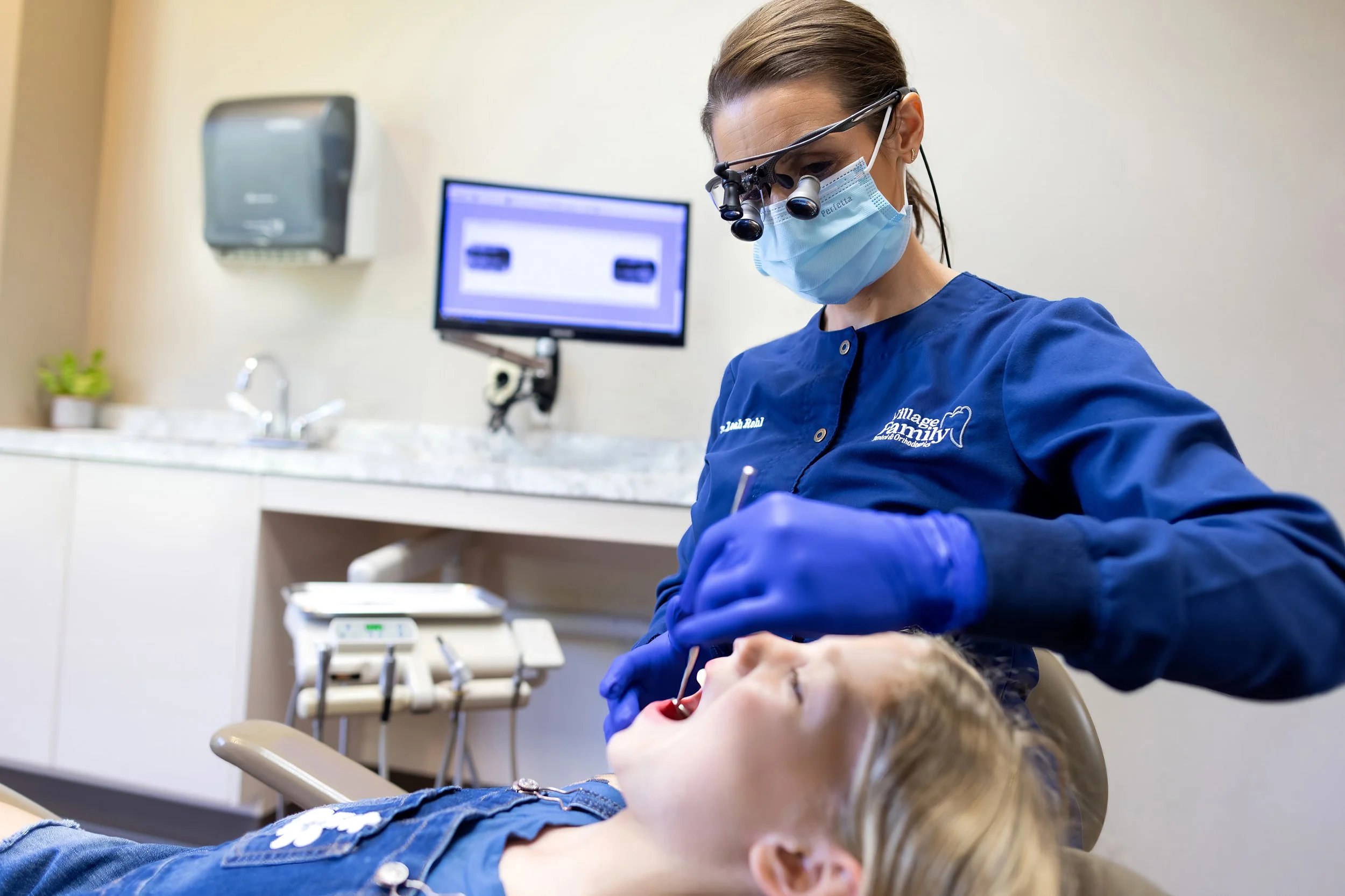 Dentist wearing magnifying glasses, mask, and gloves performing dental work on a young girl lying in a dental chair in a dental office.