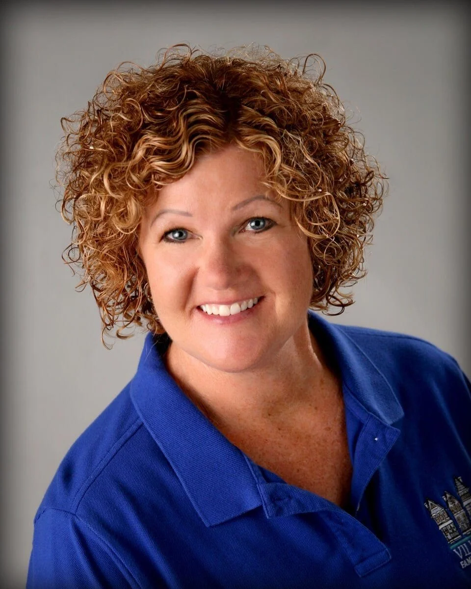 A woman with curly red hair, wearing a blue polo shirt, smiling at the camera.