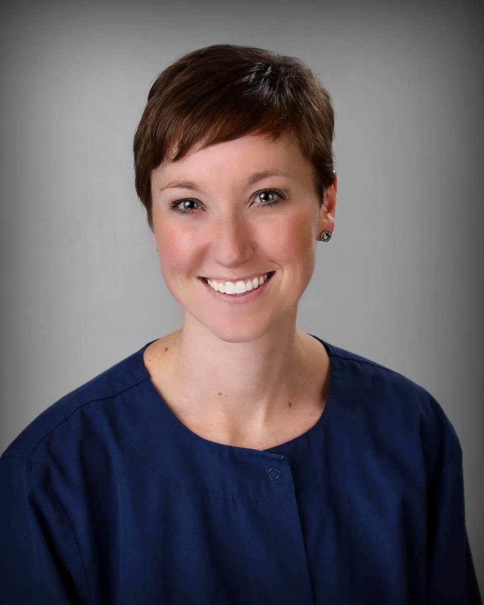 Portrait of a woman with short brown hair, wearing a dark blue blouse, smiling against a gray background.