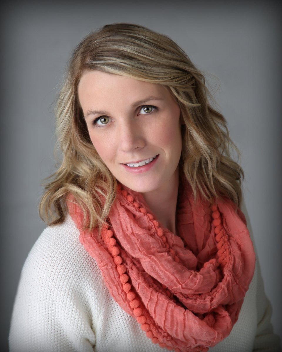 A blonde woman with wavy hair, wearing a white sweater and a coral-colored scarf, smiling at the camera against a gray background.
