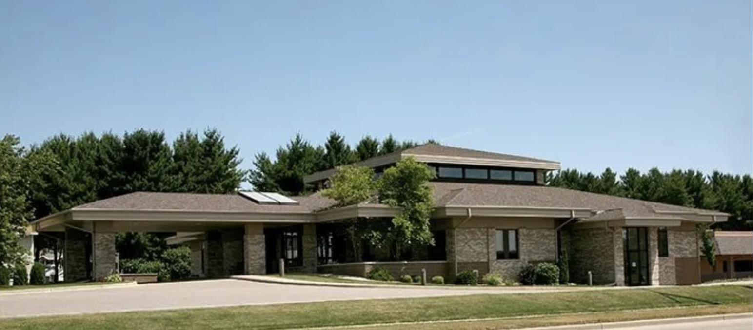 A modern office building with a sloped roof, large windows, stone and brick exterior, and solar panels, surrounded by green trees and a grassy lawn.
