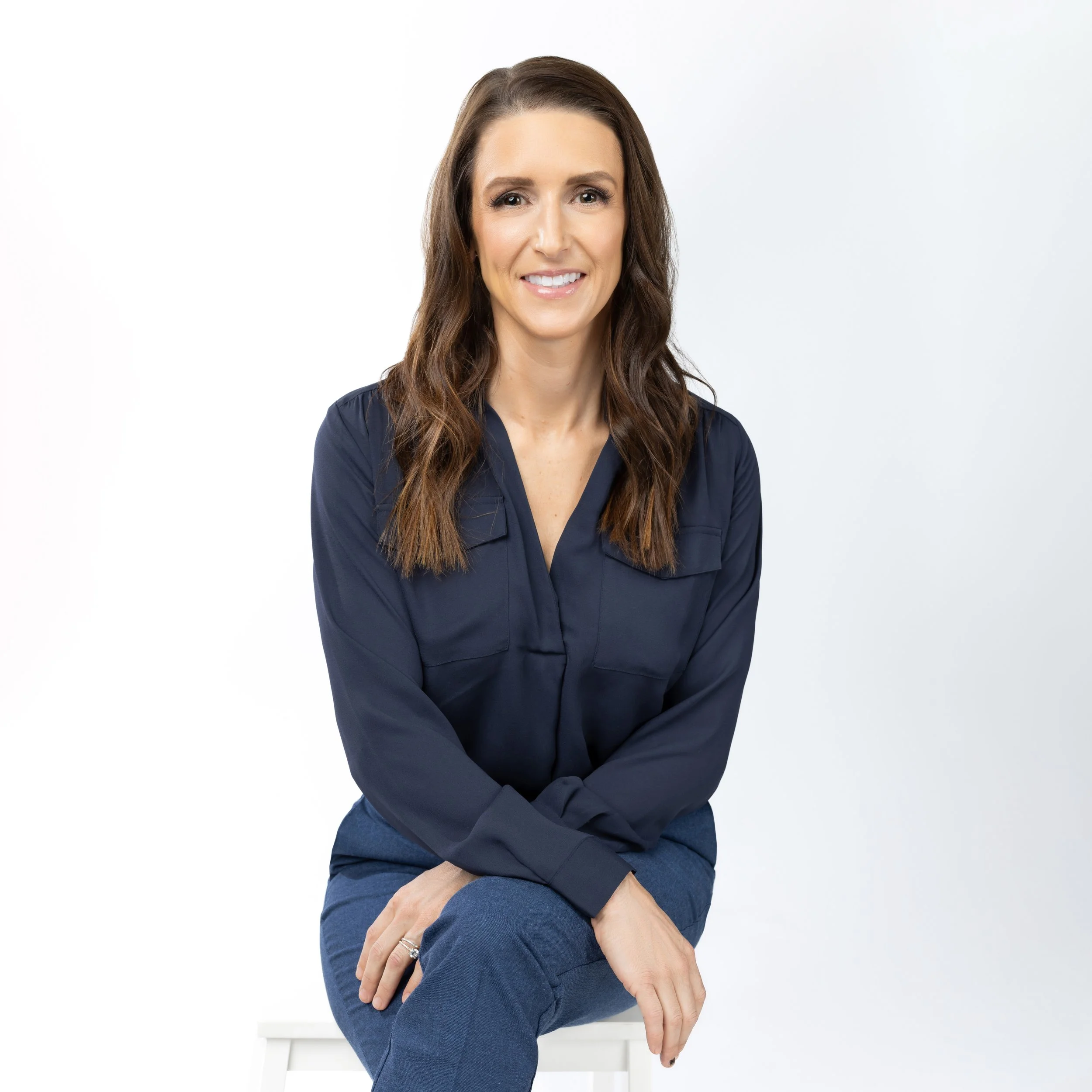 A woman with brown hair wearing a navy blue blouse and jeans, sitting on a white stool against a white background, smiling.