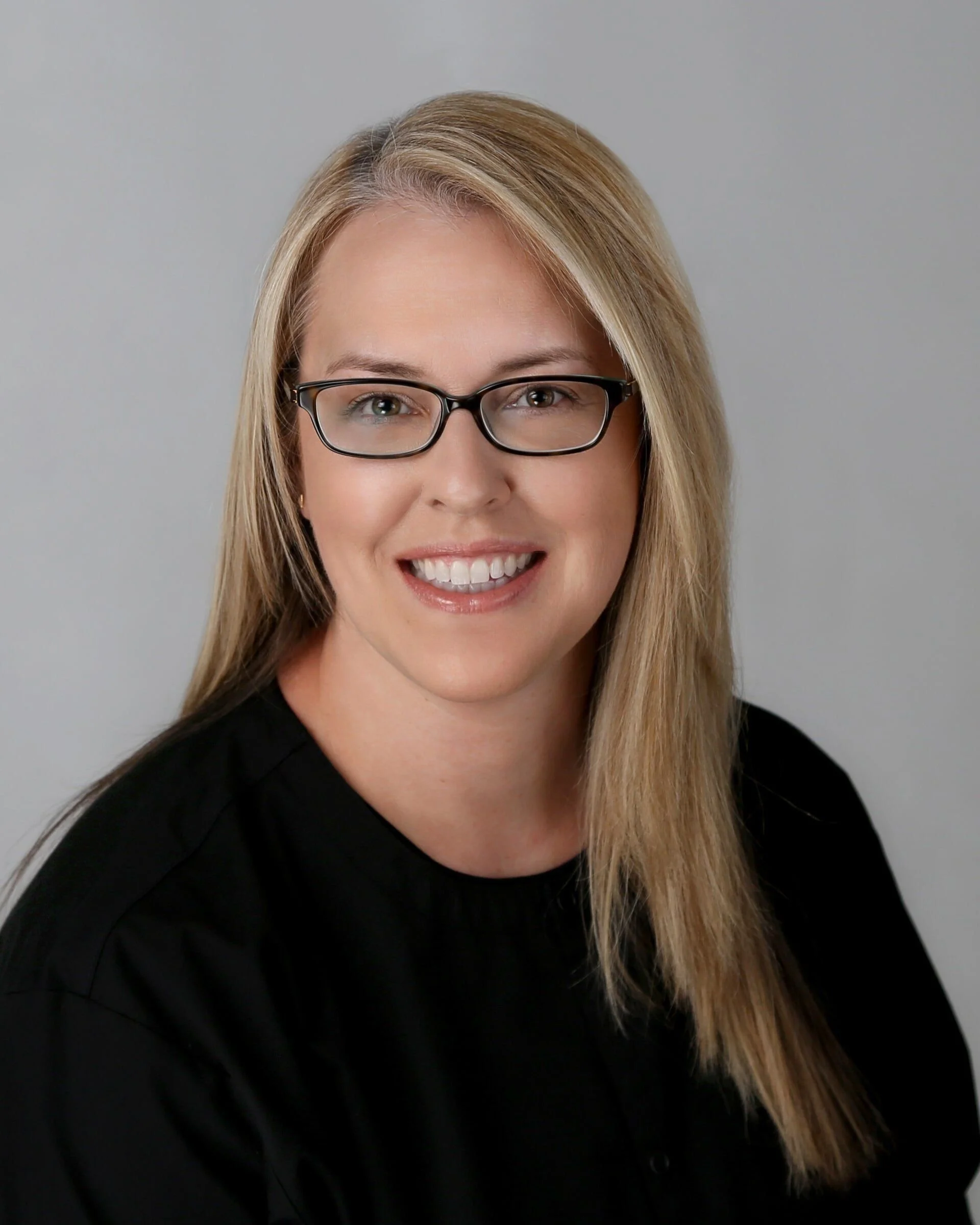 A woman with blonde hair, glasses, and a black top smiling against a plain gray background.