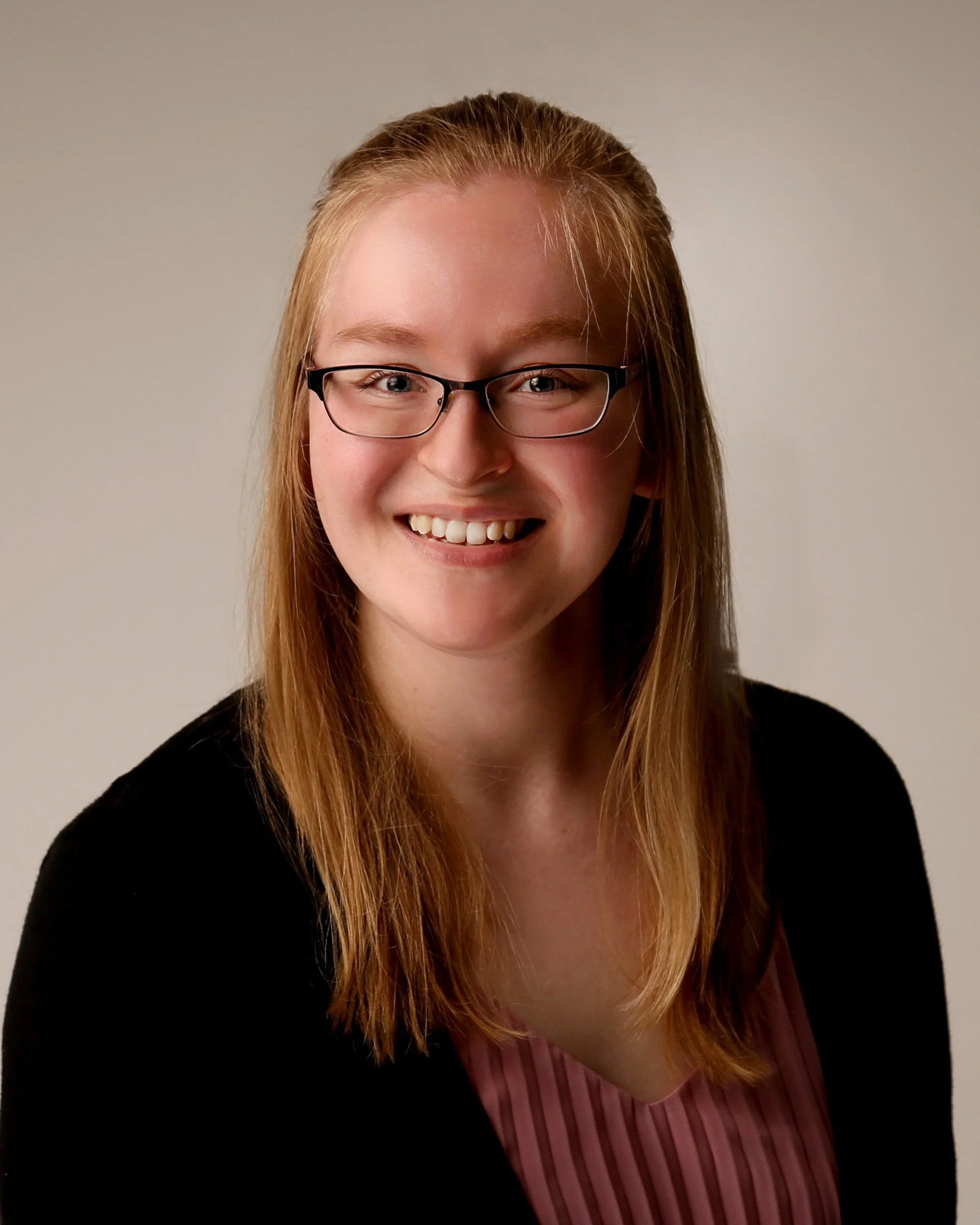 Portrait of a young woman with red hair, glasses, and a smile, wearing a black top and a pink striped blouse, against a plain background.