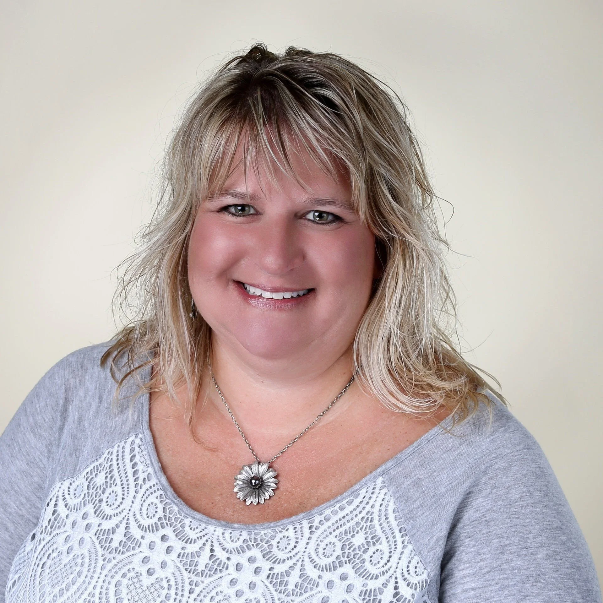 A smiling woman with blonde hair wearing a gray top with lace detail and a flower-shaped pendant necklace.