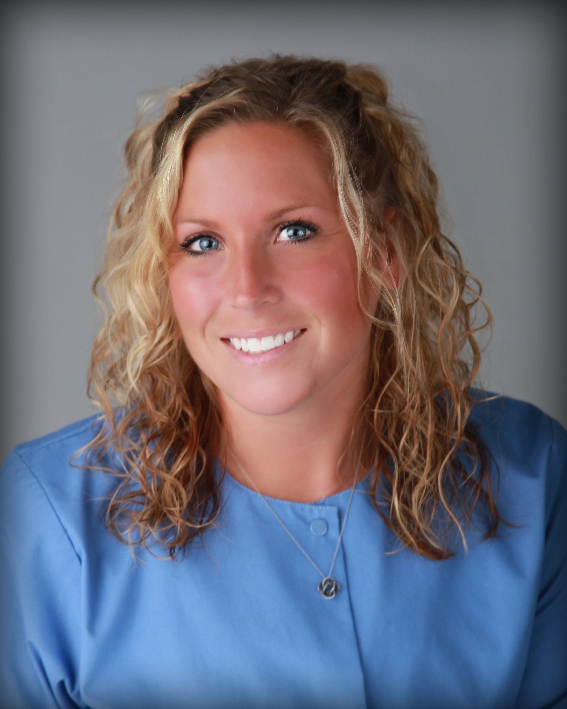 A woman with curly blonde hair and blue eyes wearing a blue medical scrubs top and a silver necklace, smiling at the camera