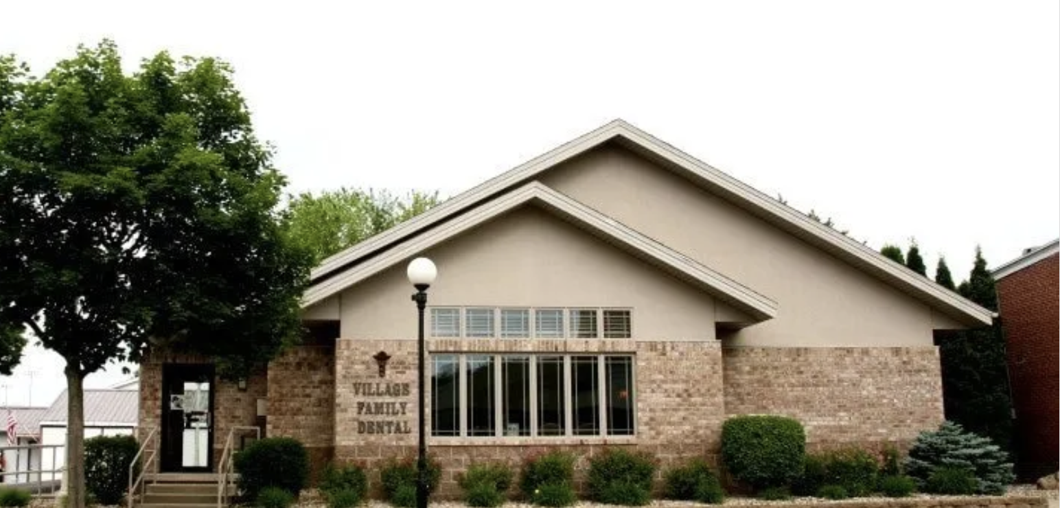 A modern brick and beige building with a sign that reads 'Villages Family Dental,' a lamp post, trees, and neatly trimmed bushes in front.