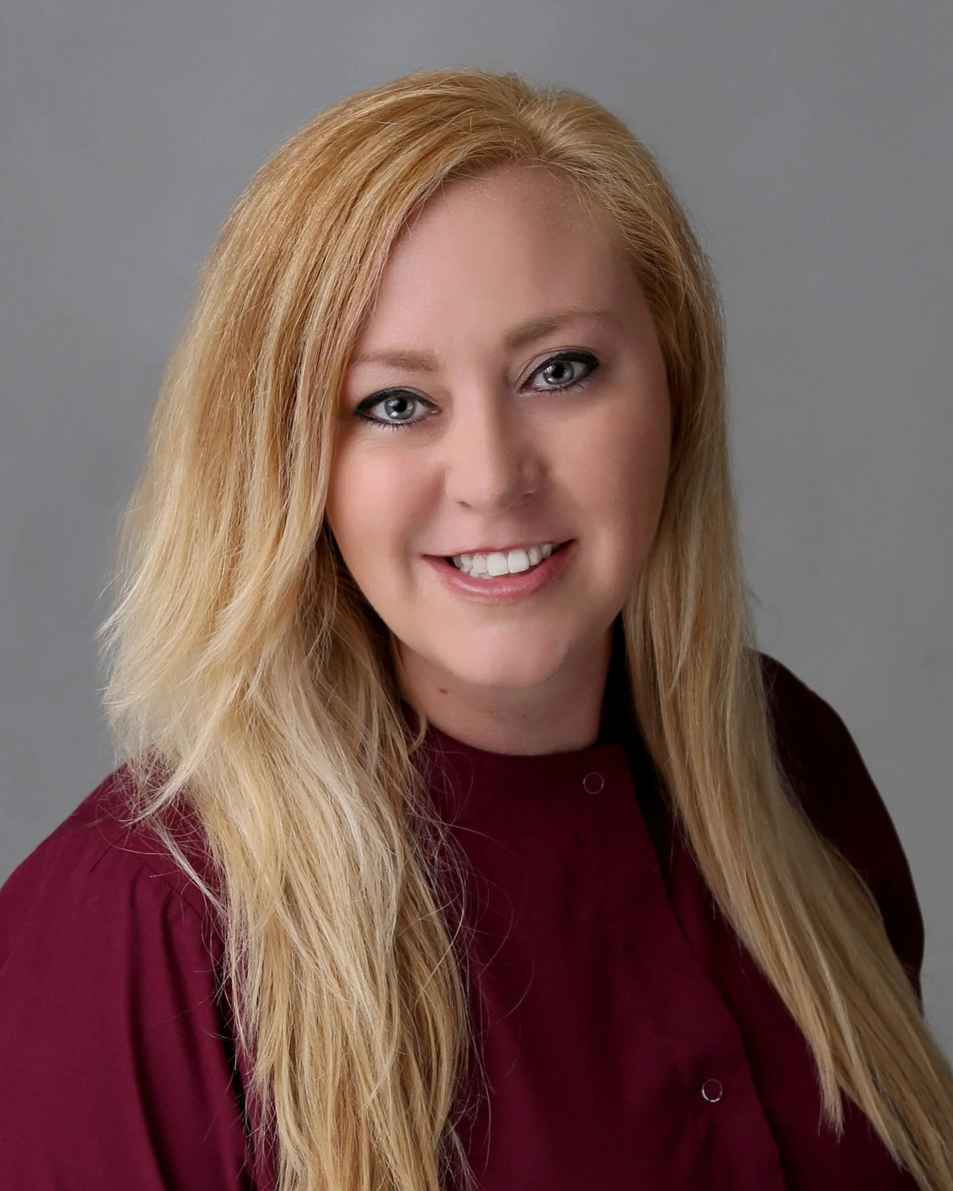 A woman with long, wavy blonde hair and blue eyes, smiling, wearing a maroon blouse, against a gray background.