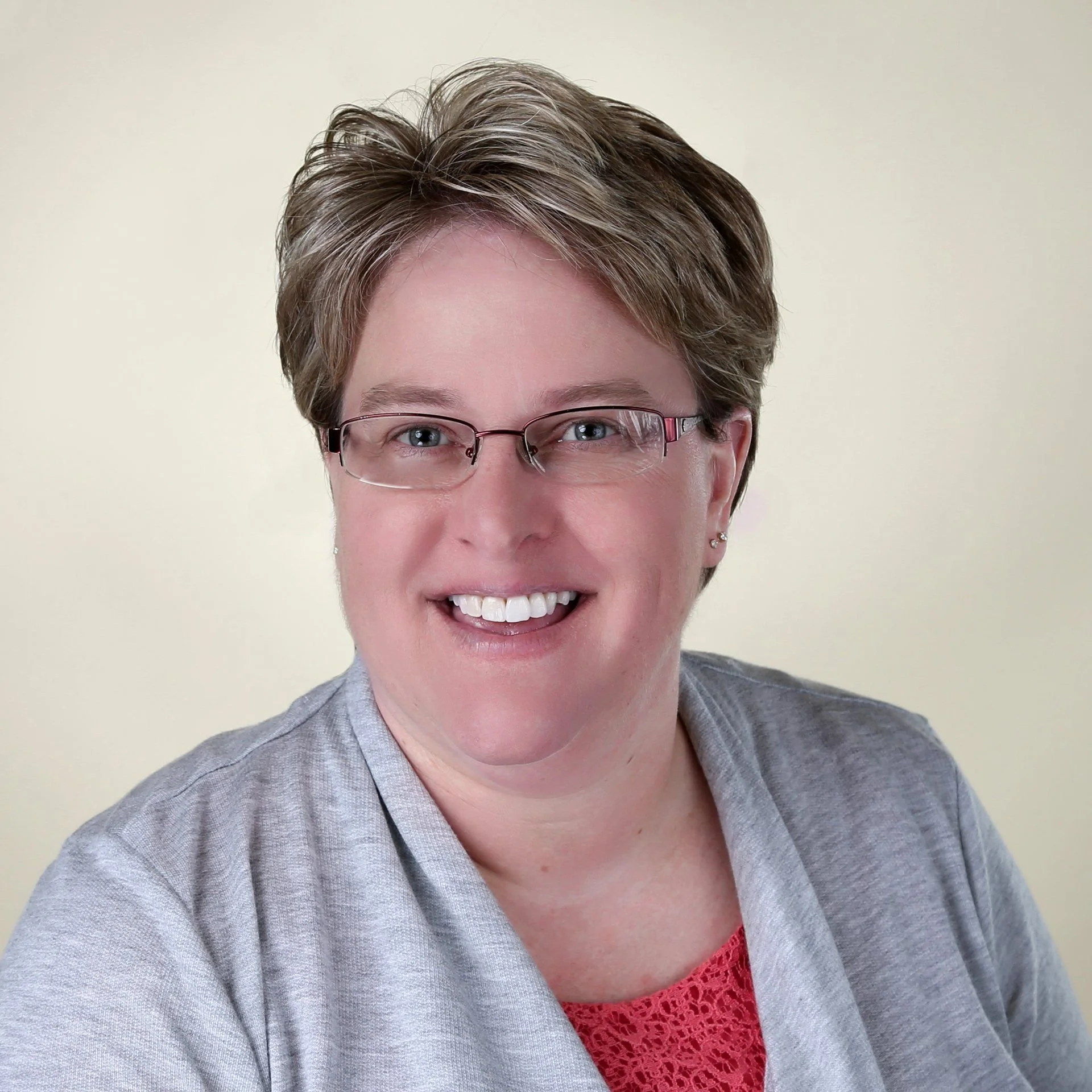 Portrait of a woman with short brown hair, glasses, wearing a gray blazer over a red top, smiling against a plain light background.