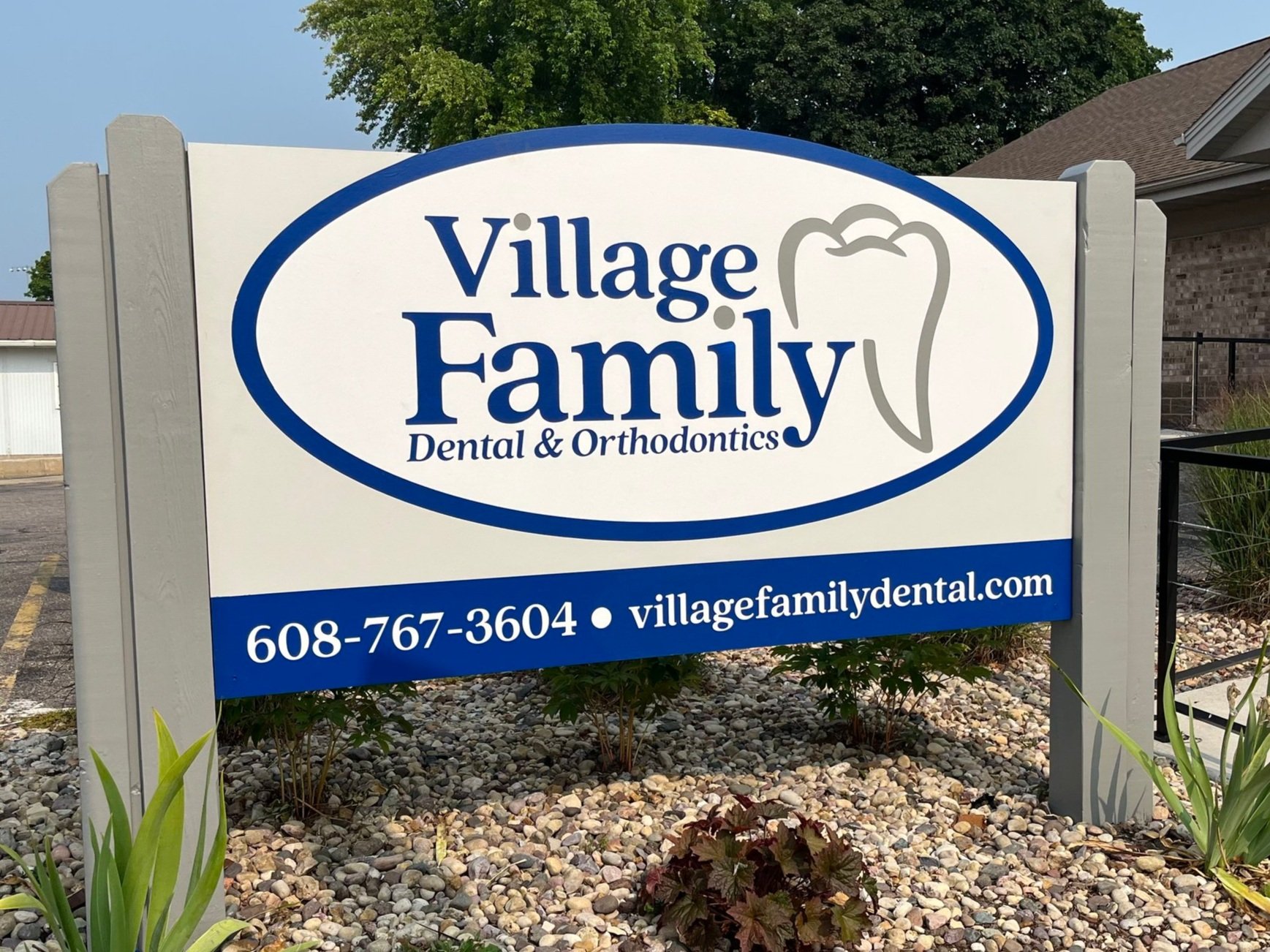 Signs for Village Family Dental & Orthodontics, with a tooth logo, phone number 608-767-3604, and website villagefamilydental.com, situated in a landscaped area with rocks and plants.