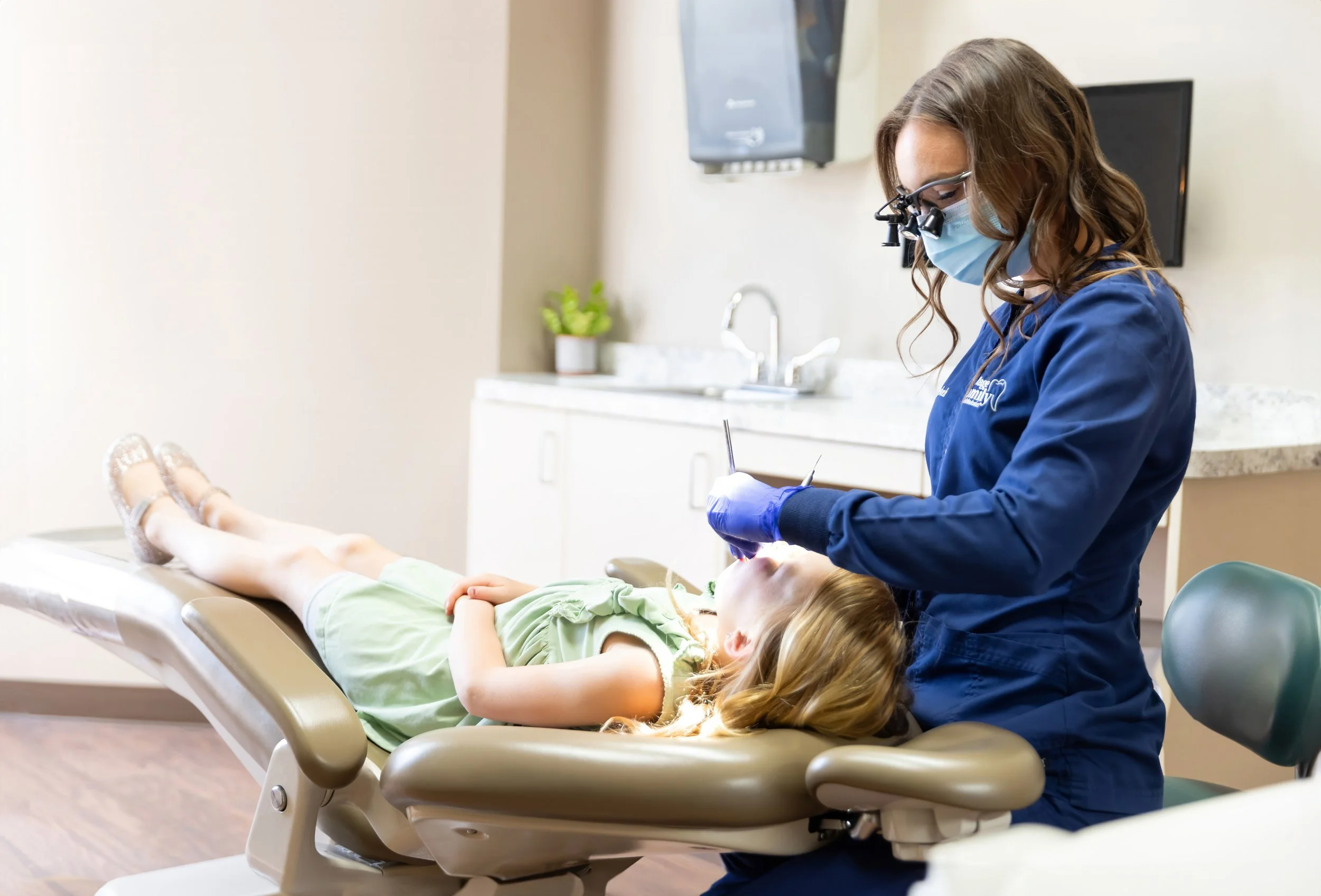 A young girl lying on a dental chair at the dentist's office, receiving dental treatment from a dentist wearing glasses, a face mask, gloves, and a blue uniform.