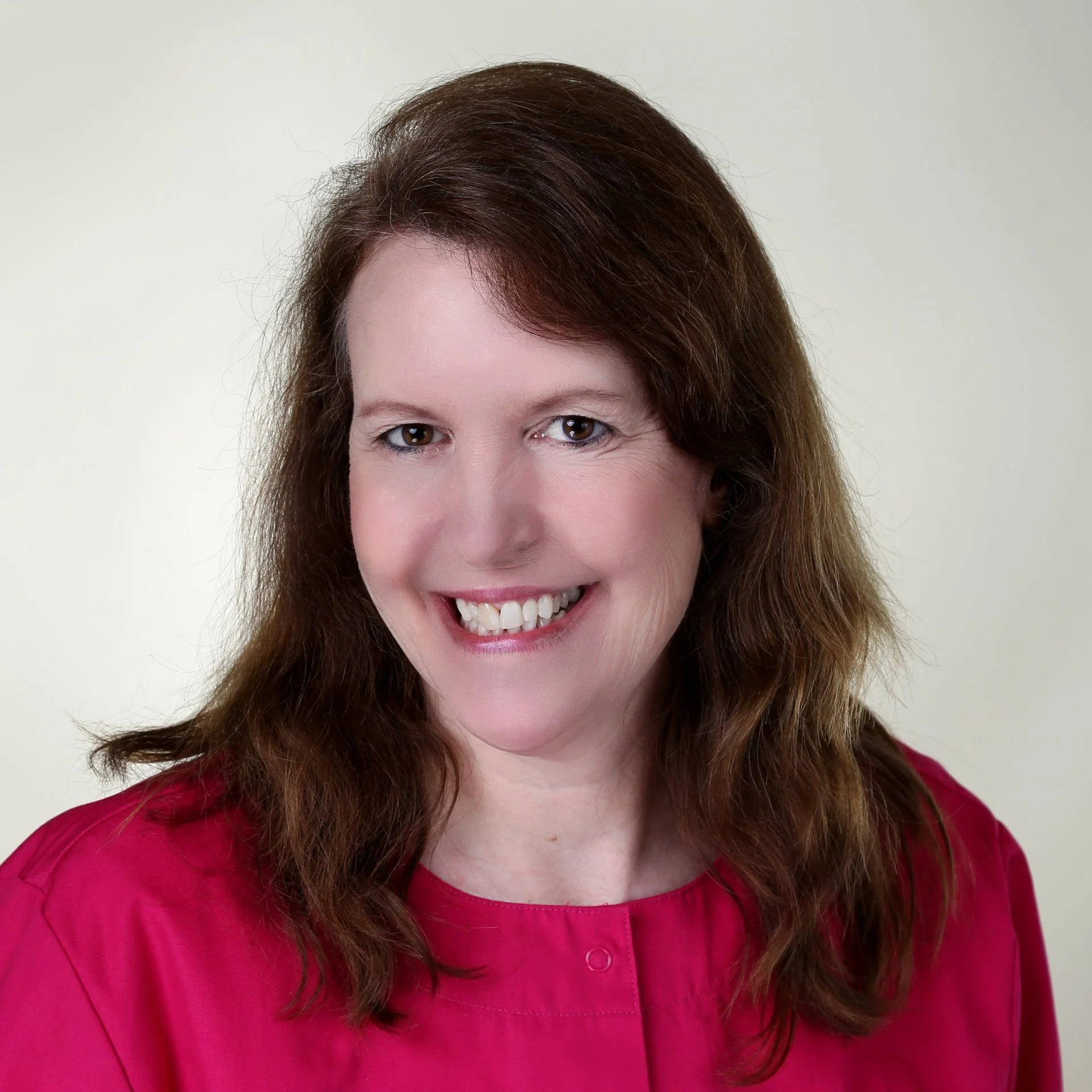 Portrait of a woman with brown hair wearing a pink top, smiling with teeth showing, against a neutral background.