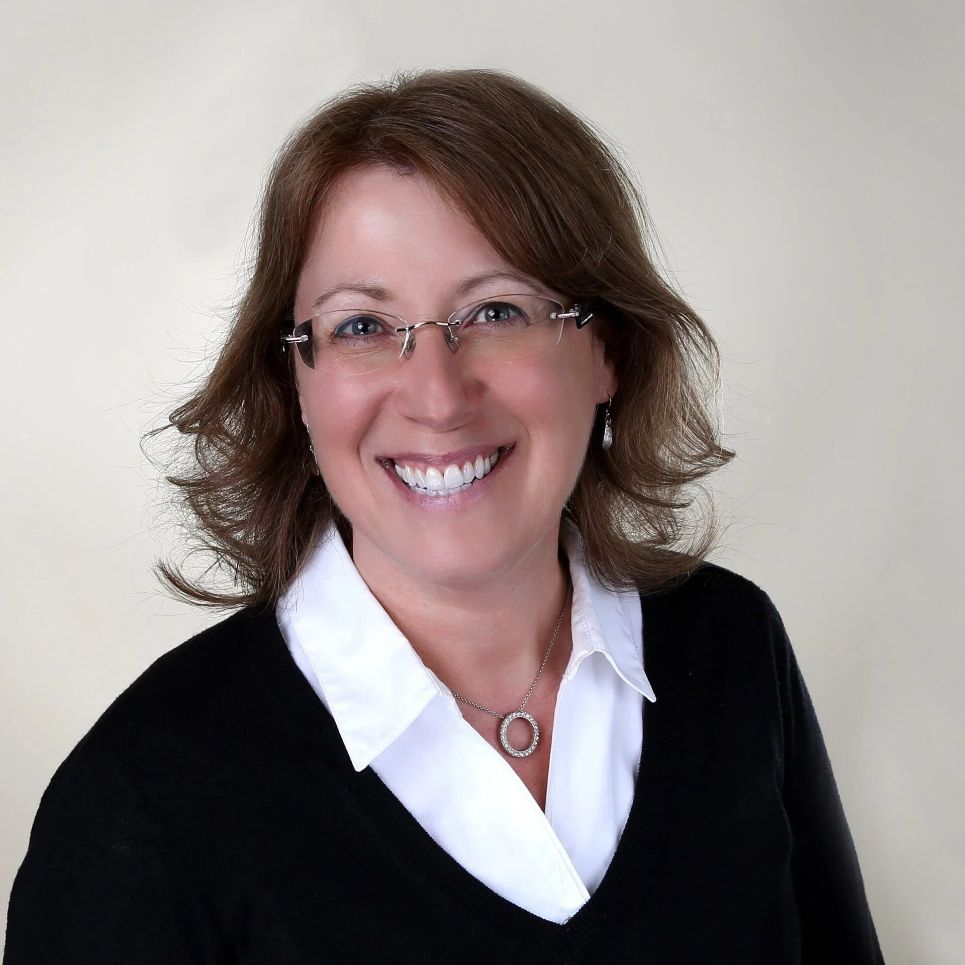 A smiling woman with light skin, shoulder-length wavy brown hair, glasses, wearing a white collared shirt, black sweater, and a silver necklace with a circular pendant, standing against a plain light-colored background.