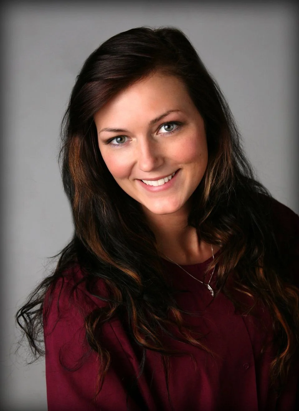A young woman with long dark hair, blue eyes, and a bright smile, wearing a maroon top and a necklace, posed against a neutral background.