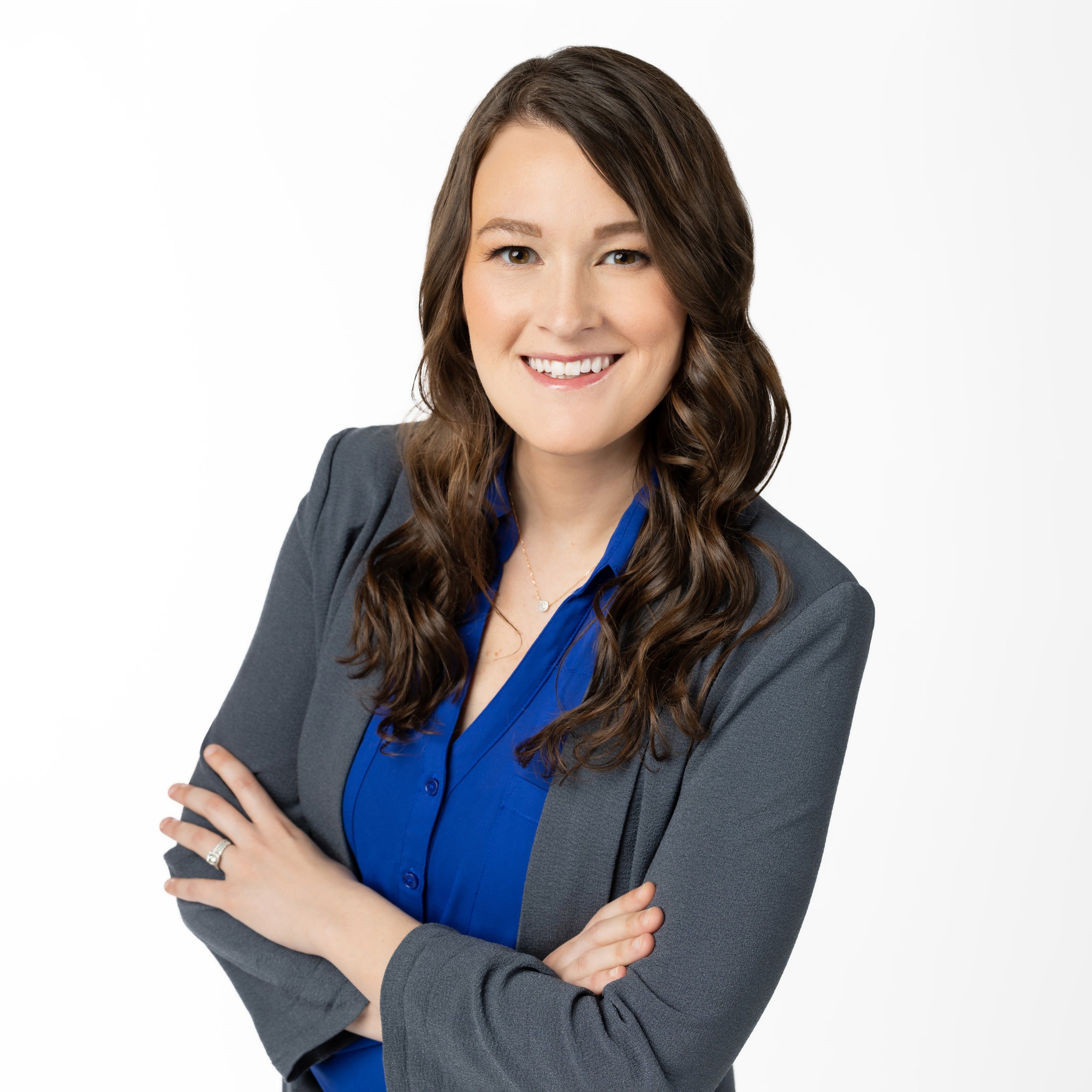 Professional woman with long brown hair in a gray blazer and blue blouse, smiling with arms crossed.