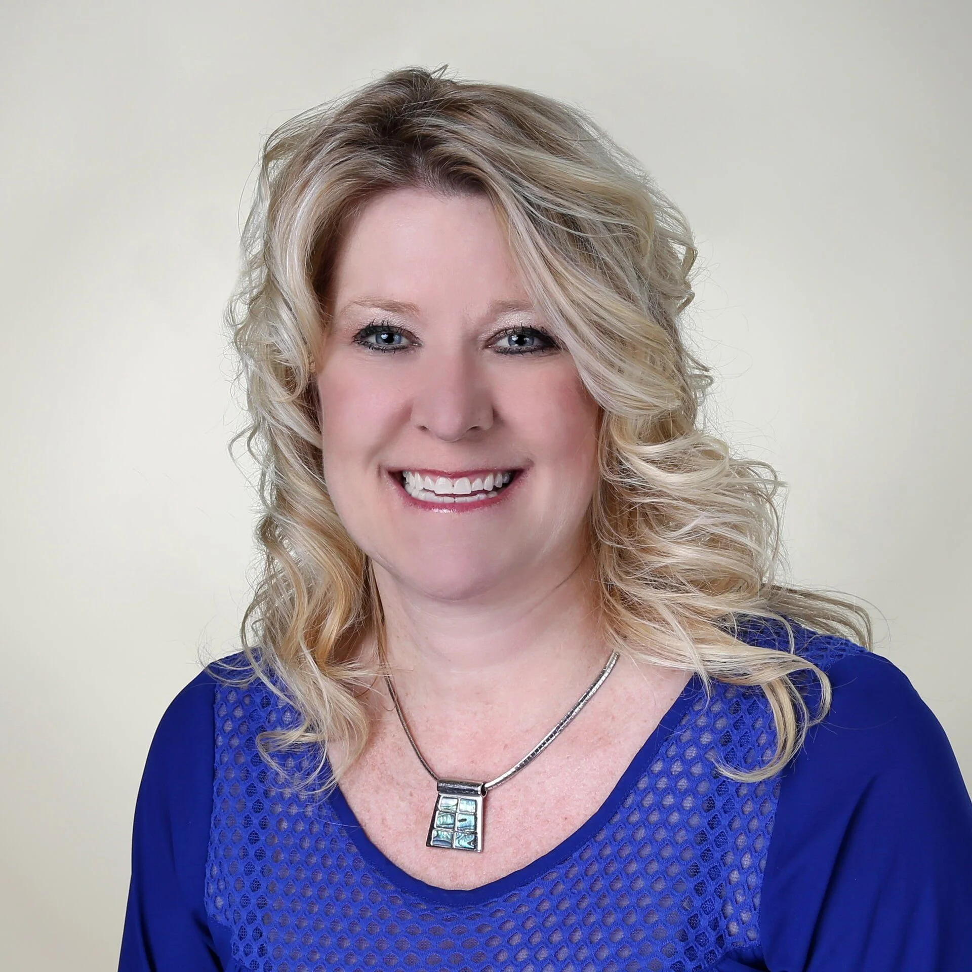 A smiling woman with blonde curly hair wearing a blue top and a silver necklace.