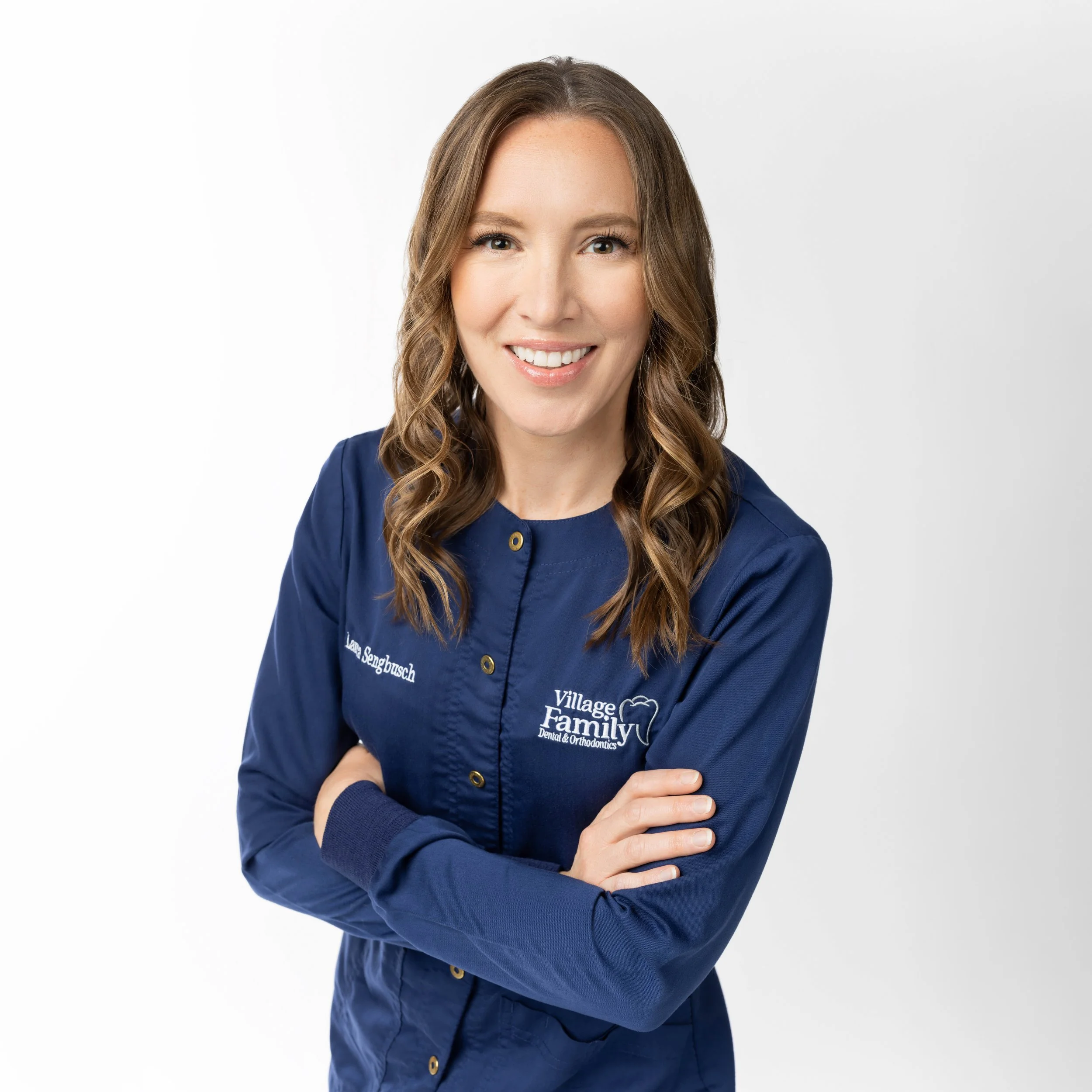 A smiling woman with long, wavy brown hair wearing a navy blue dental or orthodontic uniform with their name and logo on it, standing with arms crossed against a plain white background.