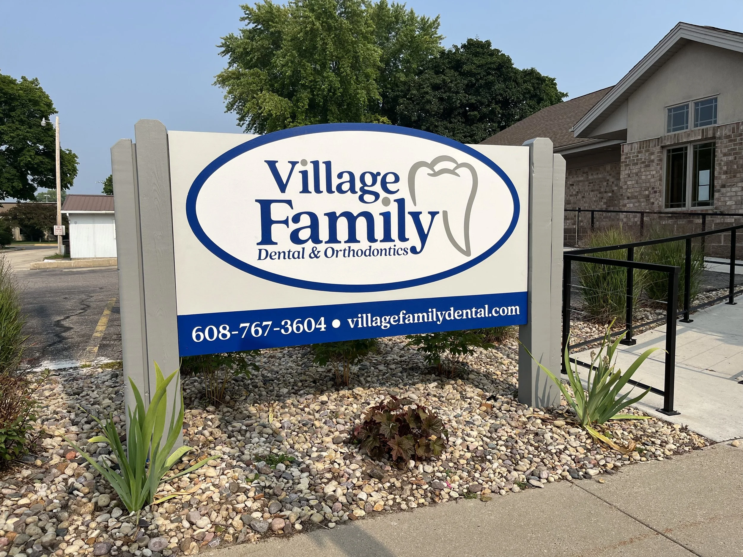 Sign for Village Family Dental & Orthodontics with a tooth logo, located in a landscaped area with rocks and plants, in front of a building with brick and siding, and a sidewalk with a ramp.