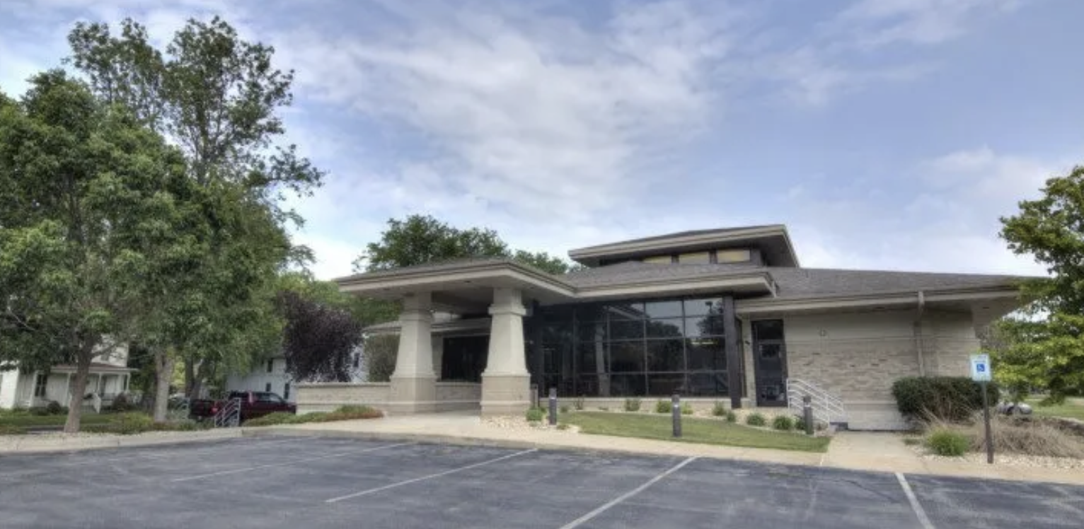 Modern commercial building with large glass windows, parking lot, and landscaped surroundings under a partly cloudy sky.