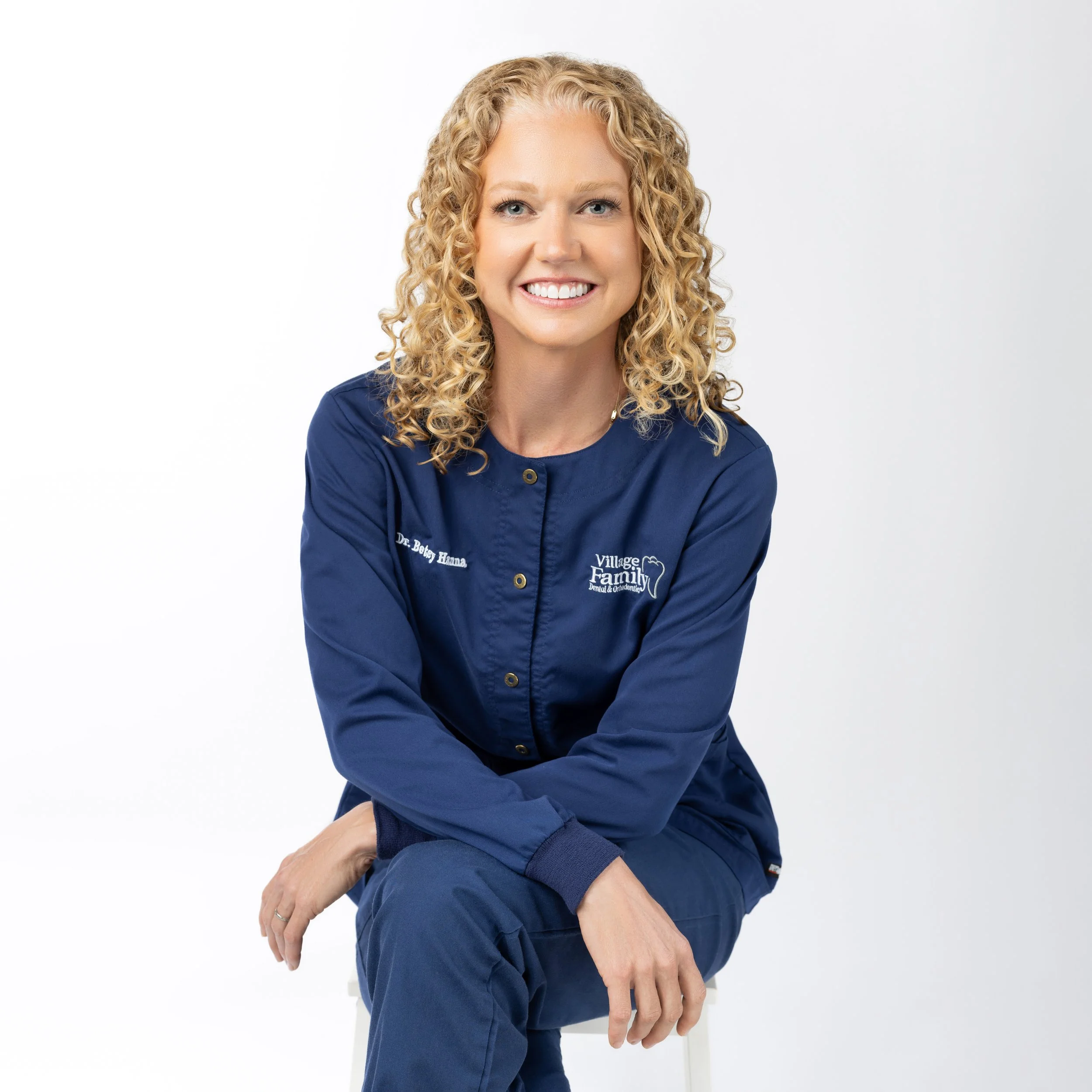 A woman with curly blonde hair smiling, wearing a navy blue dental uniform with the name tag Dr. Betsy Hanna and the logo for Village Family Dental.