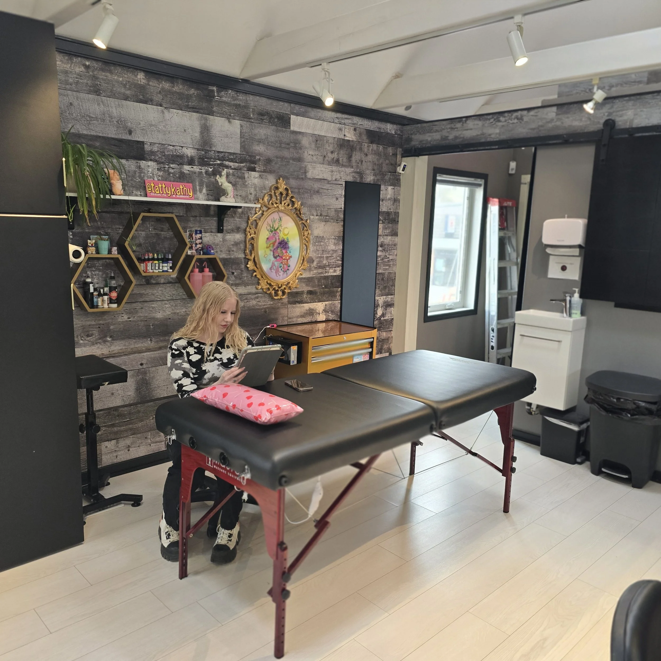 A woman with blonde hair sitting at a tattoo appointment table in a tattoo studio, with a gray wood accent wall decorated with shelves of supplies and artwork, and a painted unicorn picture on the wall.