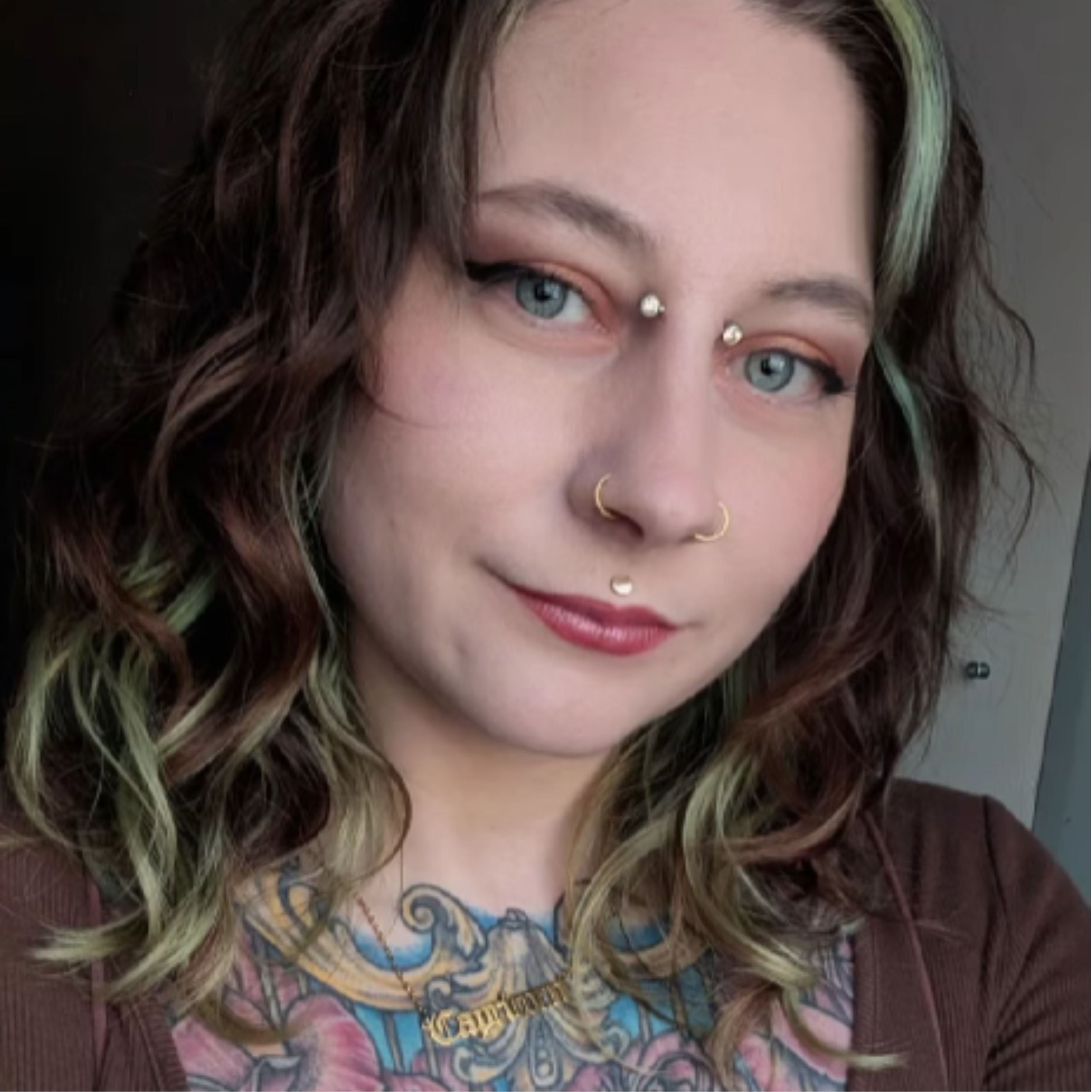 A woman with wavy brown hair with highlights, piercings on her nose and eyebrows, wearing makeup, and a tattoo on her chest, smiling softly.