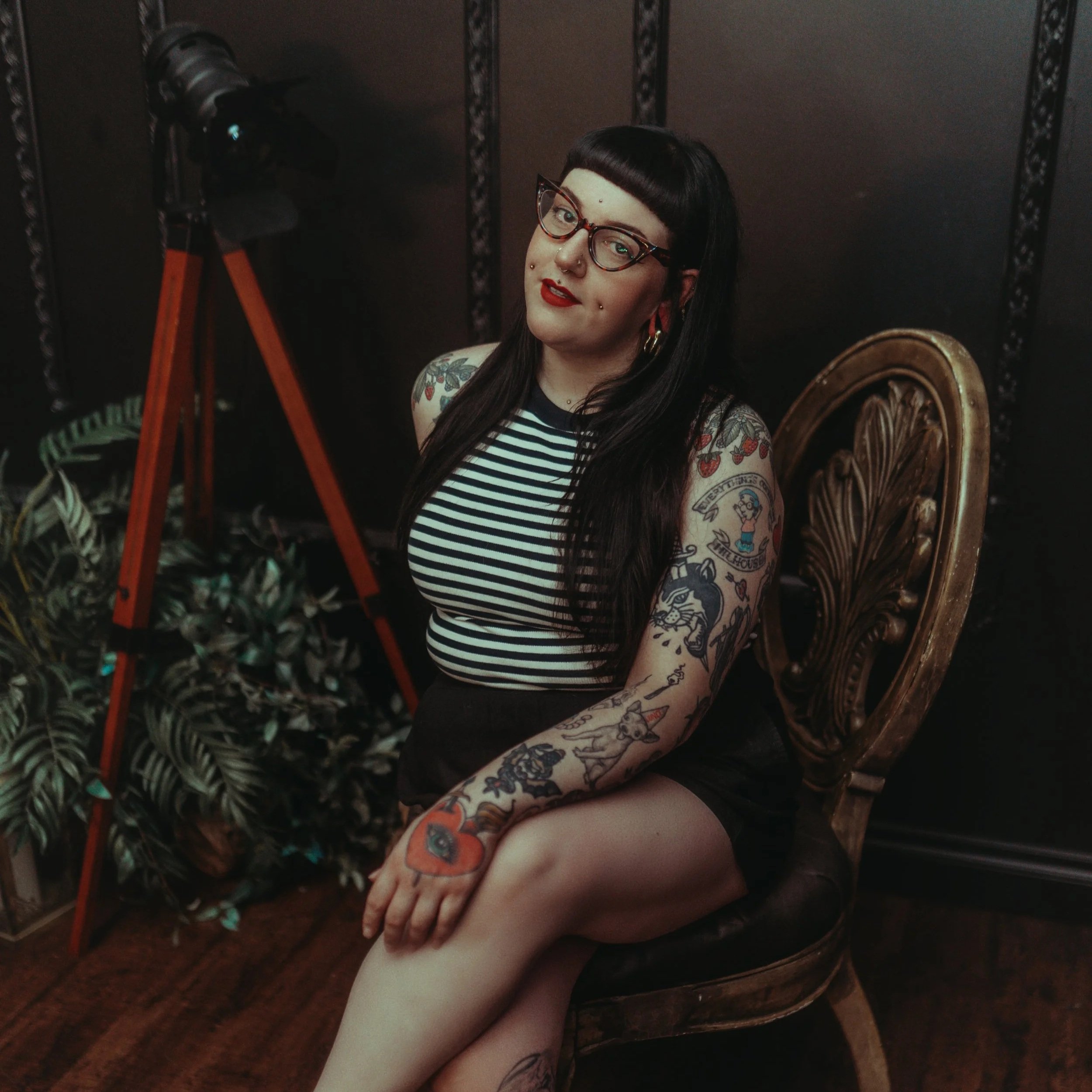 A young woman with long dark hair, glasses, and tattoos, sitting on a vintage wooden chair in a dimly lit room with plants and a large tripod camera.