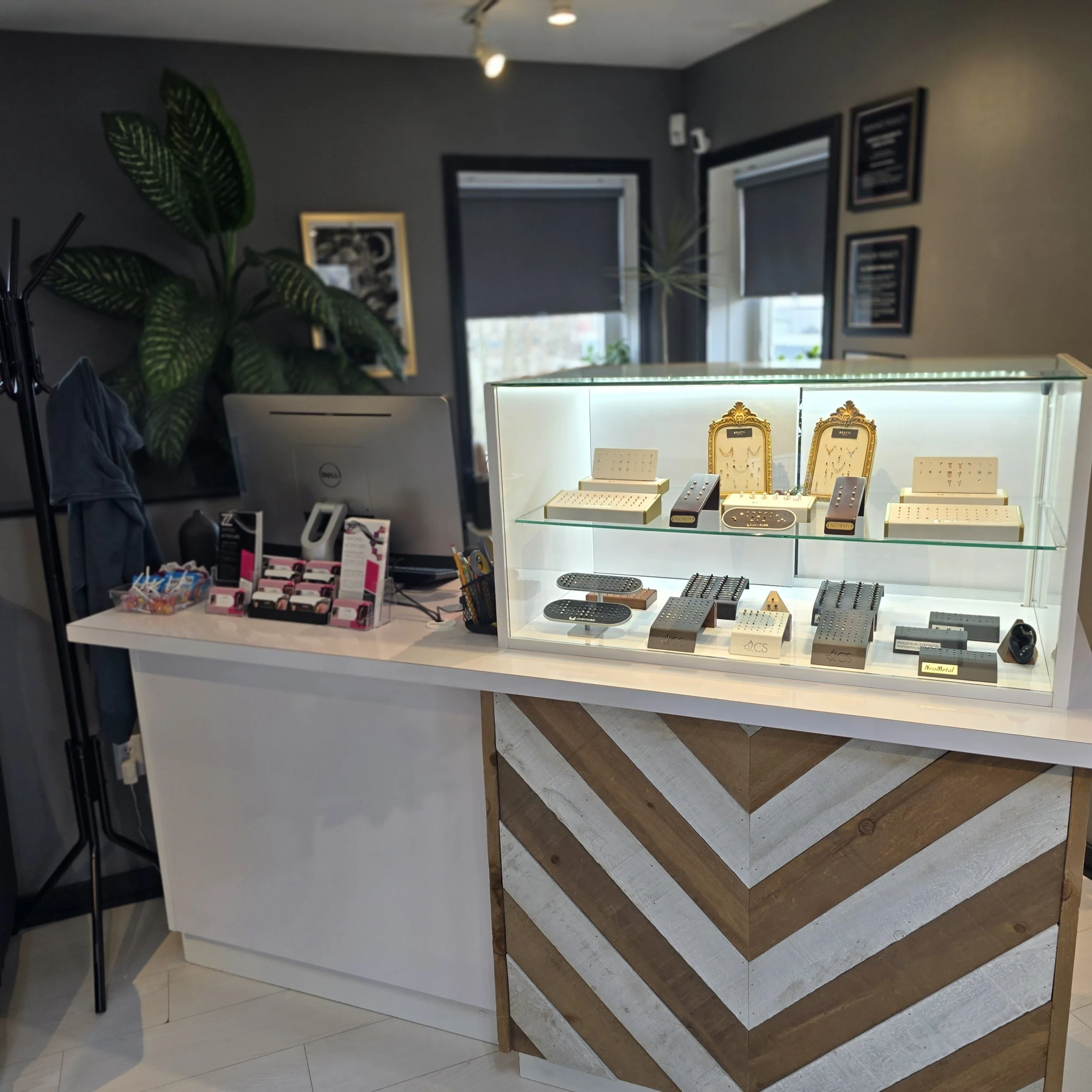 A jewelry display case with various pieces of jewelry, including rings, necklaces, and earrings, on a white surface, in a store setting with framed pictures on the wall behind and a large plant nearby.
