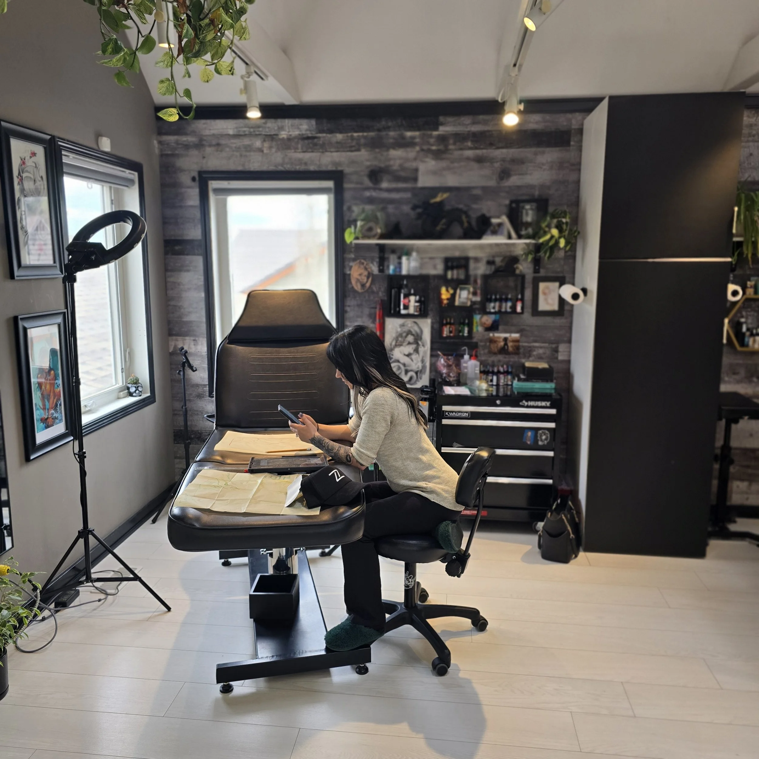 A woman with long dark hair, wearing a beige sweater and green slippers, is sitting at a black desk with her head down, looking at her phone. The room has a gray and black color scheme with wood-paneled walls, a window, and shelves with tools and decorations. A salon chair is visible behind her, and a ring light with a green plant is attached to the wall.