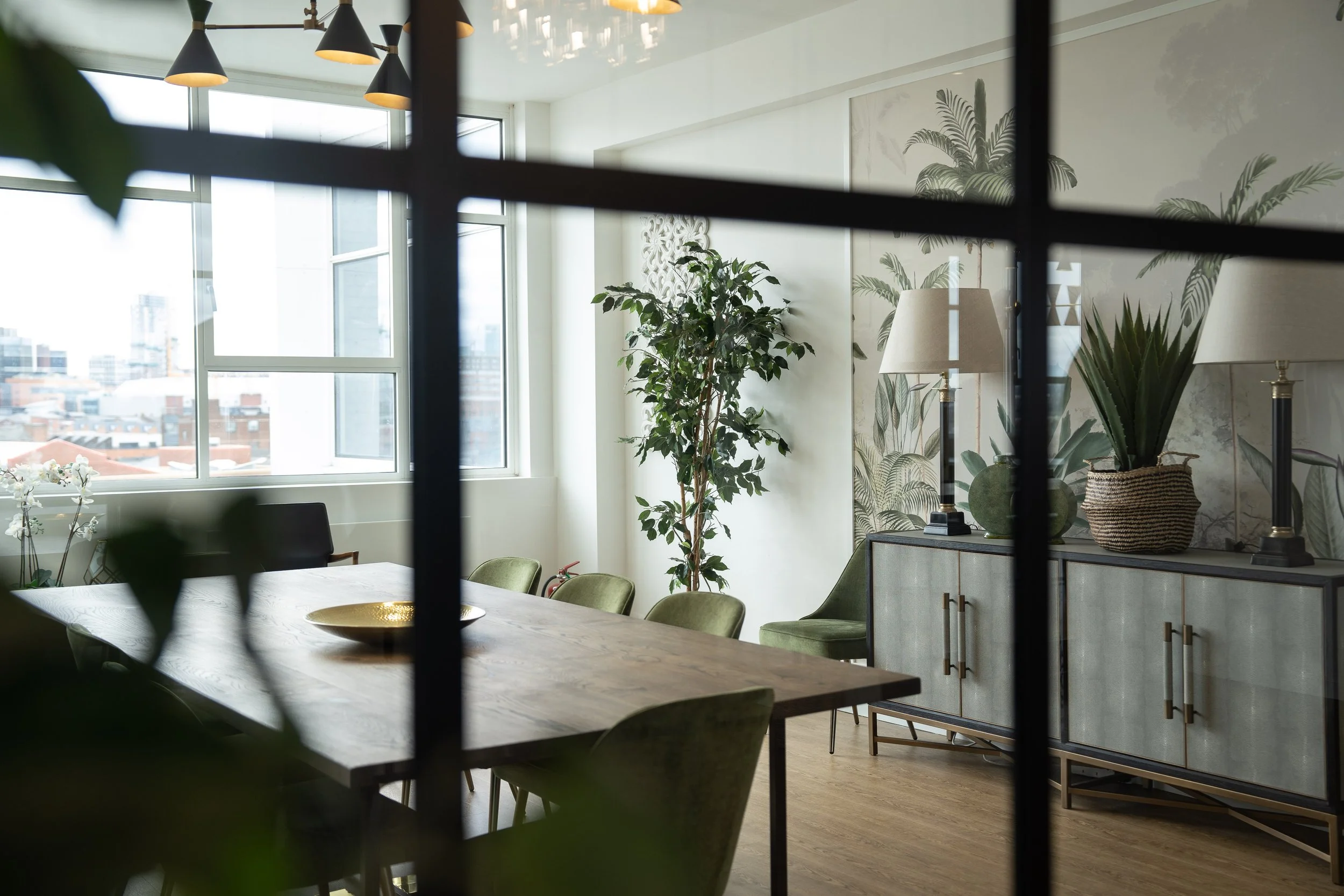 Modern dining room with large window, wooden table, green chairs, plants, and a sideboard with lamps and decor, seen through a black grid.
