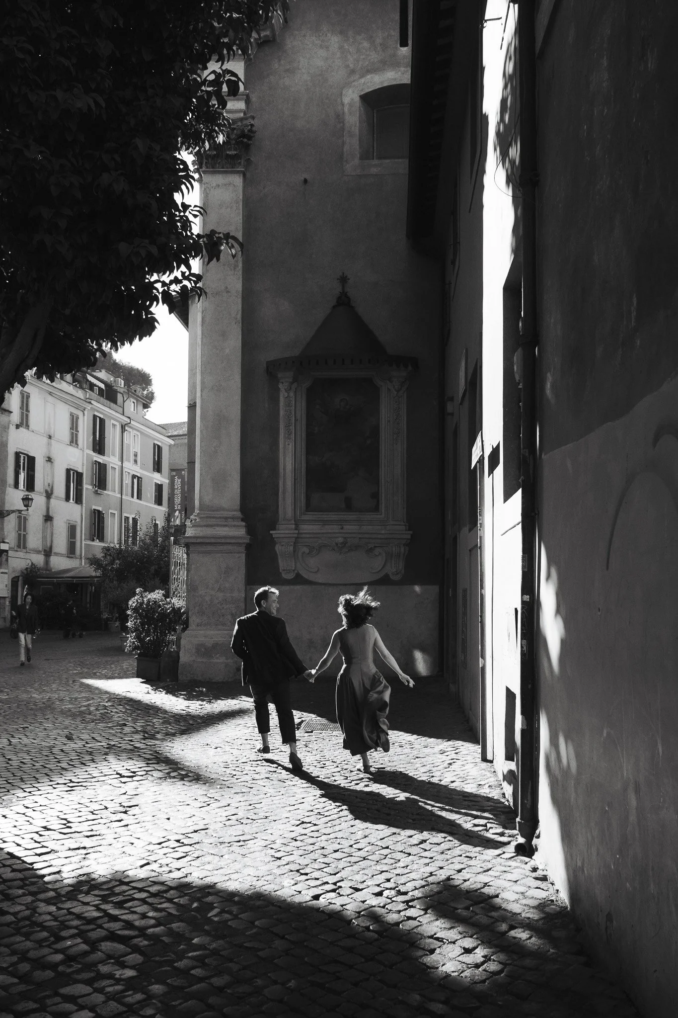 Couple photograph taken in Rome, capturing two people walking hand in hand through a narrow historic street in strong afternoon light. The black and white image focuses on movement, shadow, architecture and a spontaneous sense of intimacy within the 