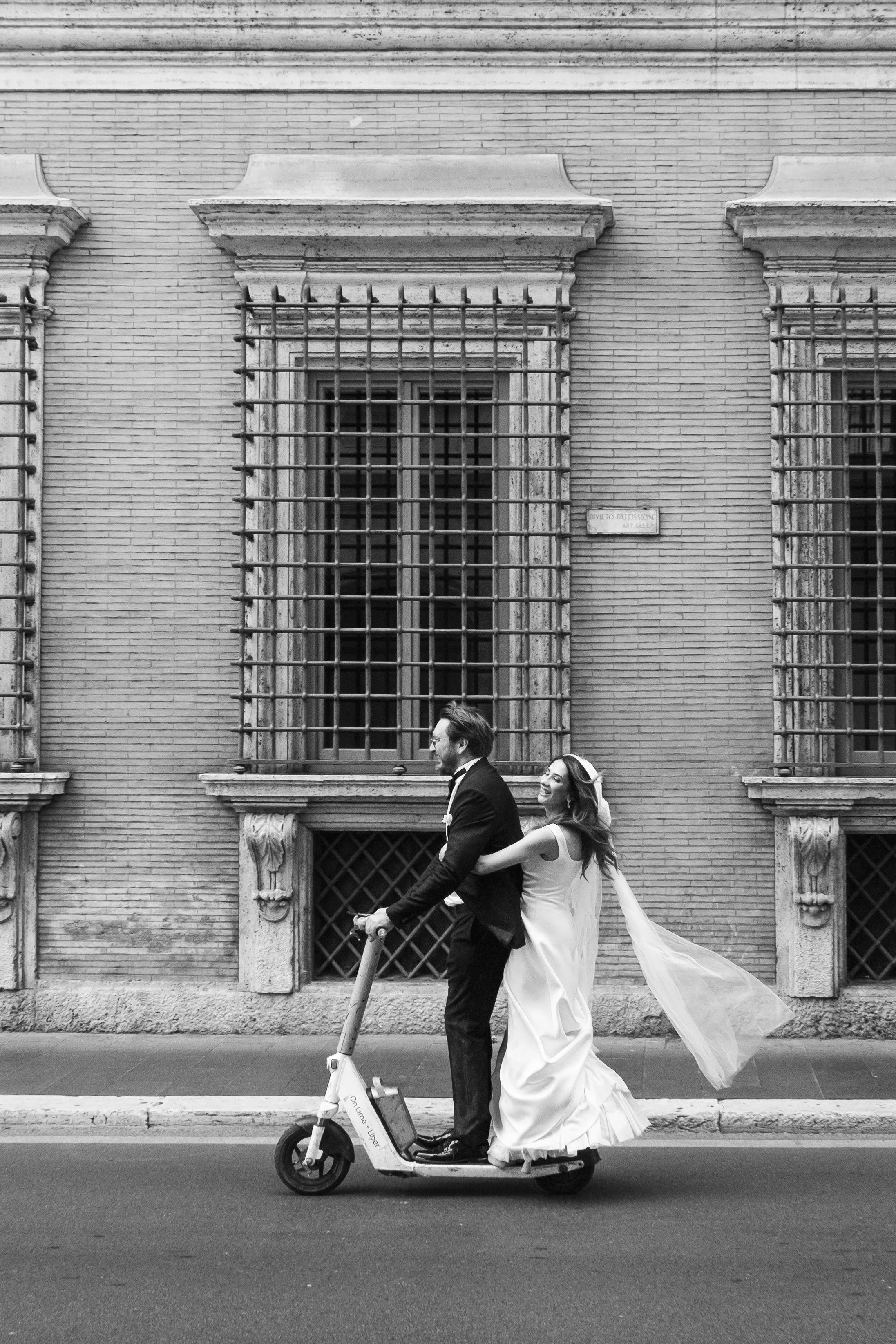 Elopement wedding photograph taken in Rome, capturing a newlywed couple riding together on an electric scooter through the city. The black and white image combines spontaneity, movement and the architecture of Rome, reflecting the playful and unconve
