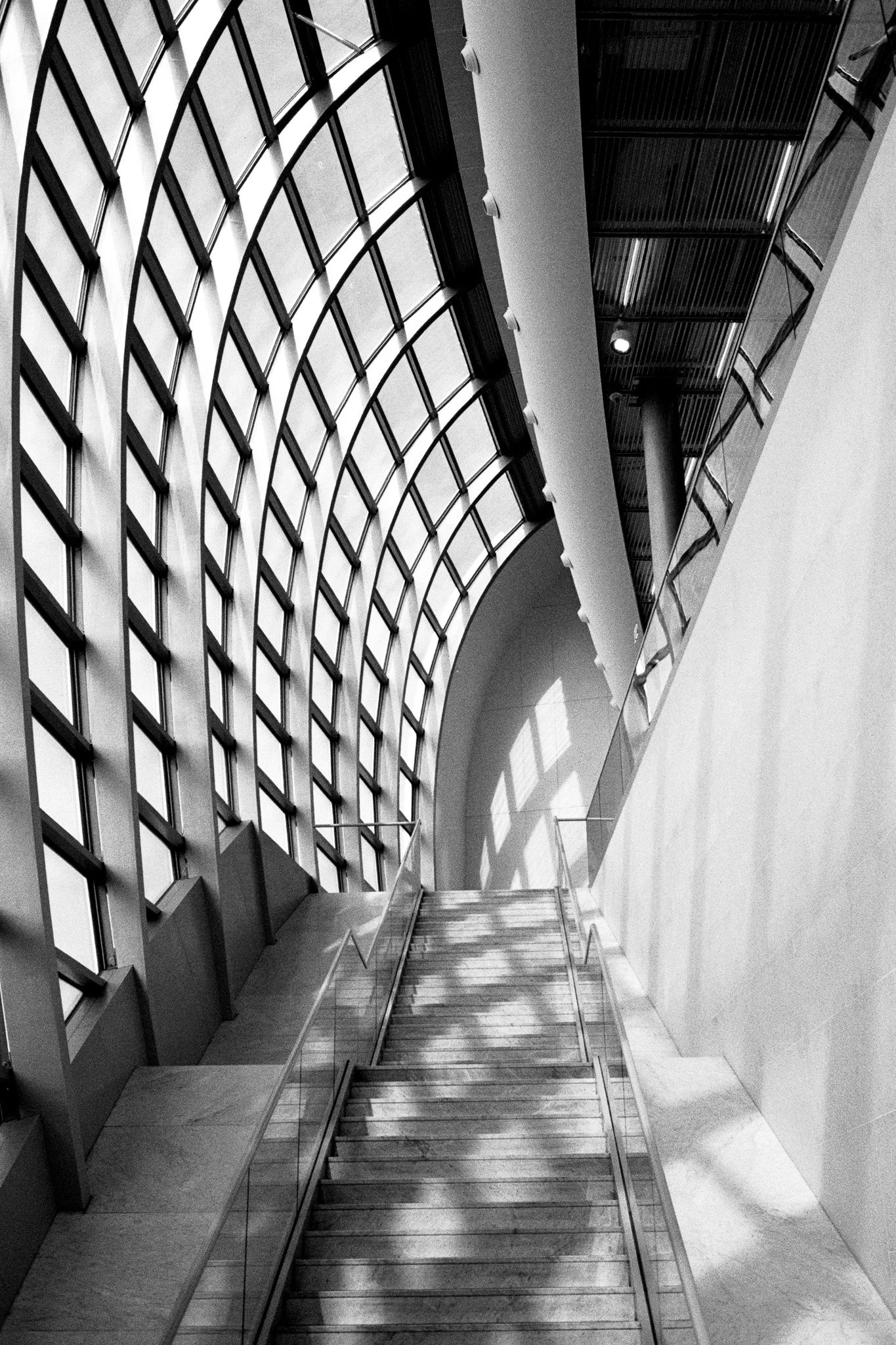 A black and white architectural photograph taken inside Paris Charles de Gaulle Airport, focusing on geometric lines, natural light, stairs and strong graphic shadows. Photography by Gökhan Kutluer.