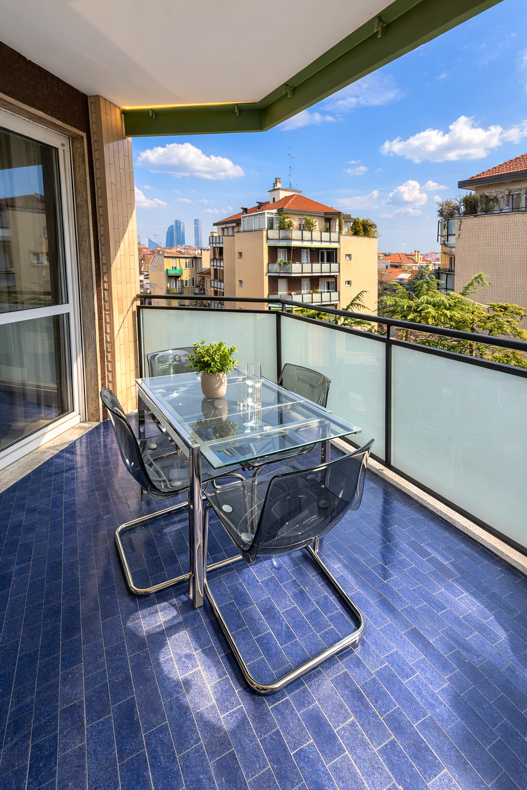 Balcone di un appartamento con tavolo in vetro, quattro sedie di plastica trasparente e un vaso con piante verdi. Vista su altri edifici e cielo azzurro con nuvole.