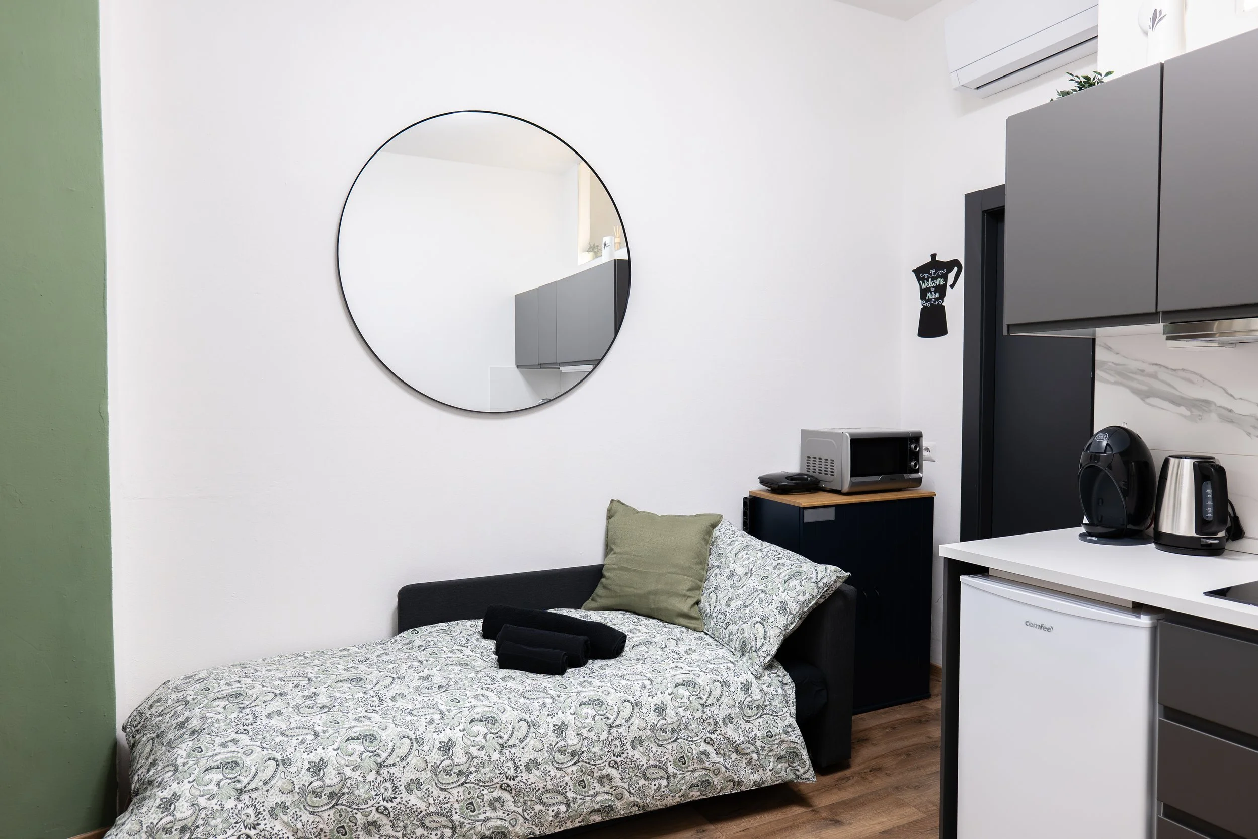 Camera piccola con letto matrimoniale, cuscini verdi e patterned, specchio rotondo sulla parete bianca, elettrodomestici neri e acciaio nella cucina moderna con armadietti grigi, frigorifero e forno.