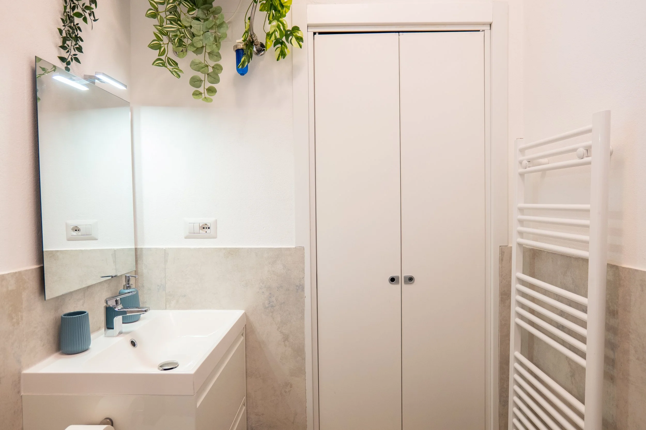 Bagno con lavabo bianco, specchio, asciugamano riscaldato e armadio bianco, piante appese al soffitto.