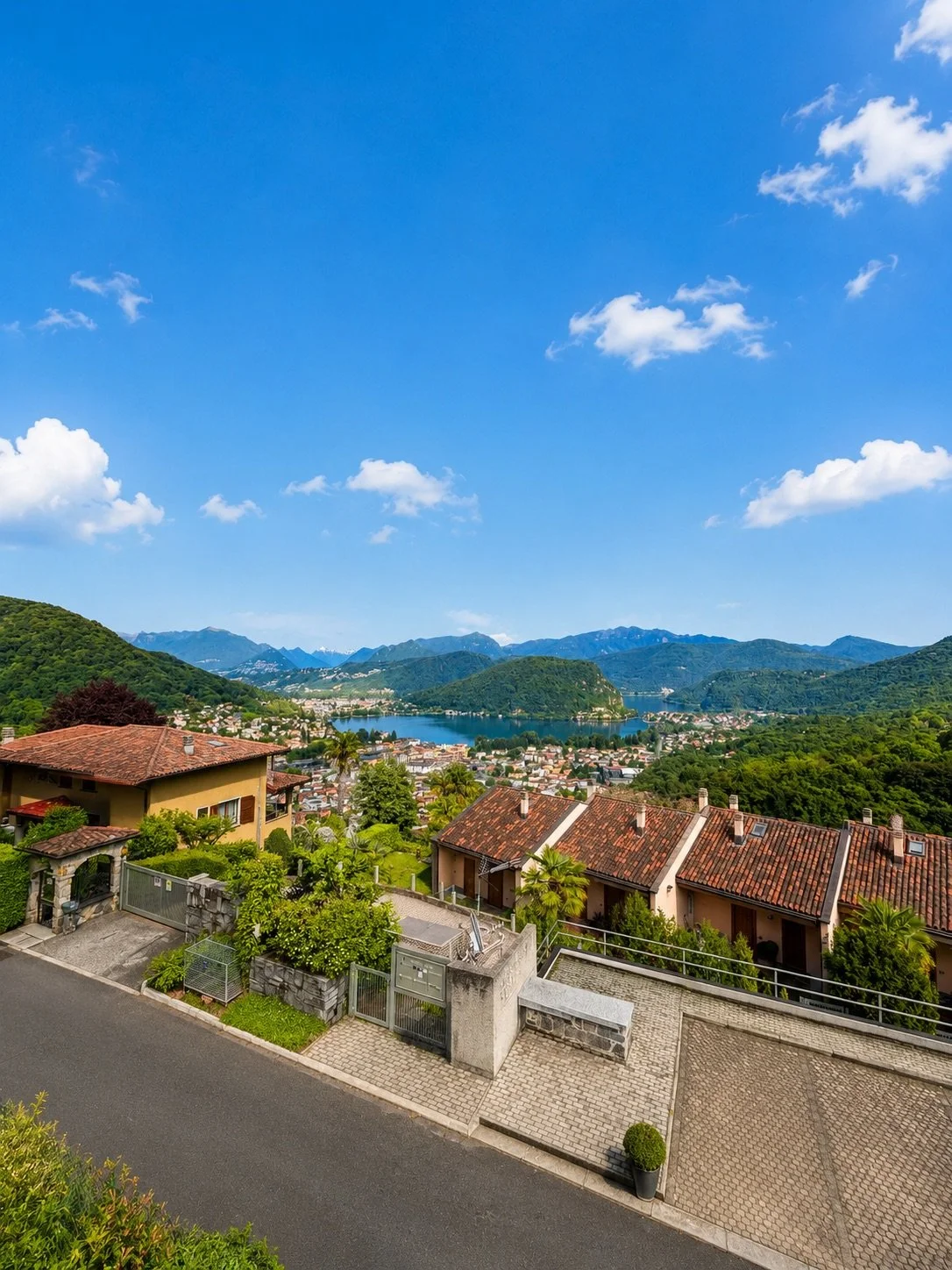 Vista panoramica di case con tetti in terracotta, un lago circondato da montagne verdi e un cielo azzurro con alcune nuvole.
