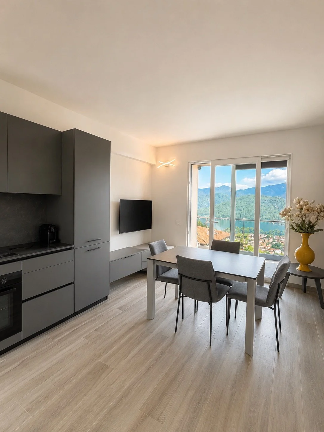 Interno di un soggiorno con cucina moderna, tavolo da pranzo con 6 sedie, grande finestra con vista sul lago e sulle montagne, decorazioni con vaso di fiori giallo.
