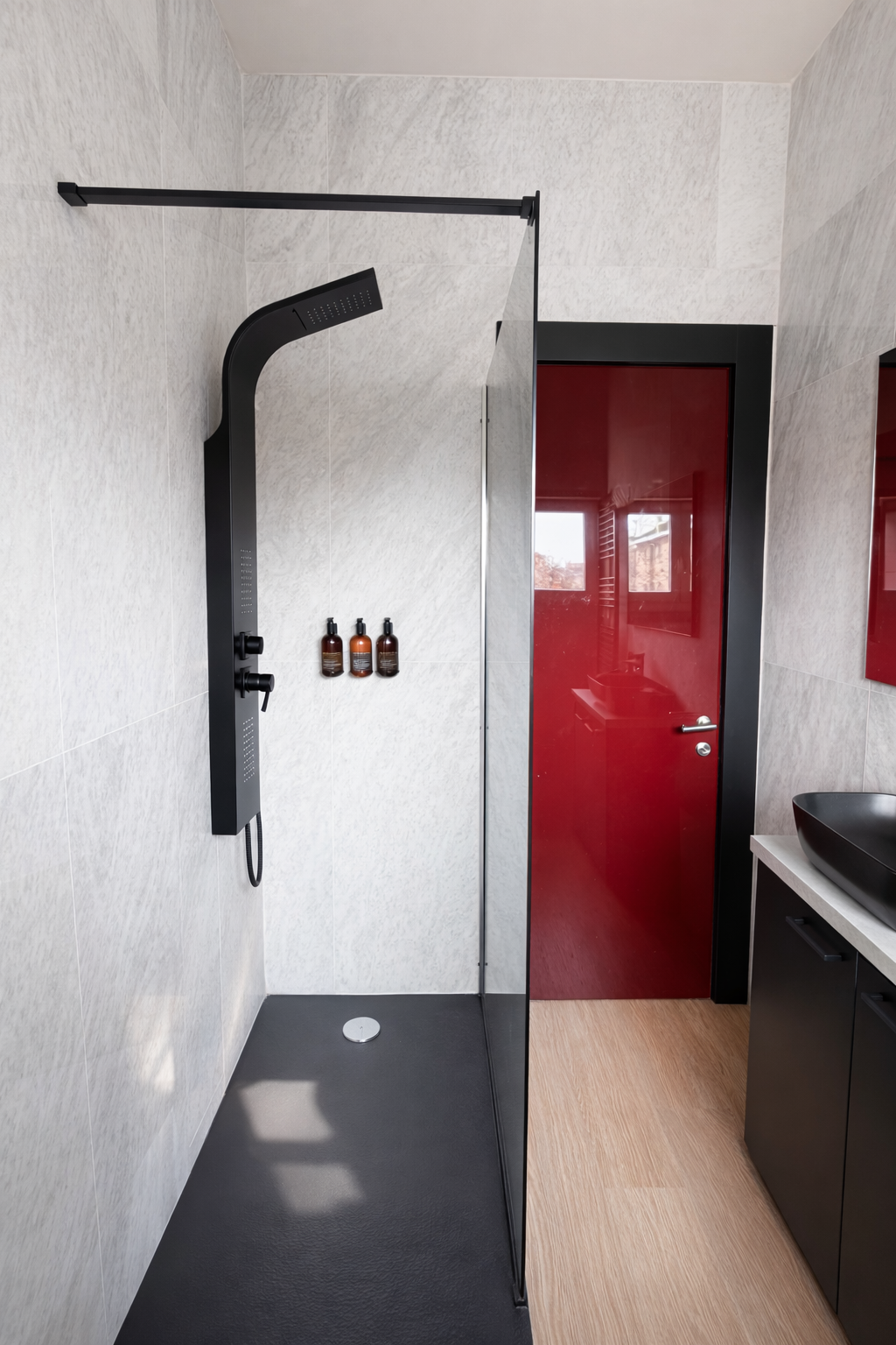 Doccia a soffione nero con pareti in piastrelle chiare, tre bottiglie di prodotti per la cura personale sulla parete, porta rossa in metallo, lavabo nero con mobile sotto, pavimento in legno chiaro.