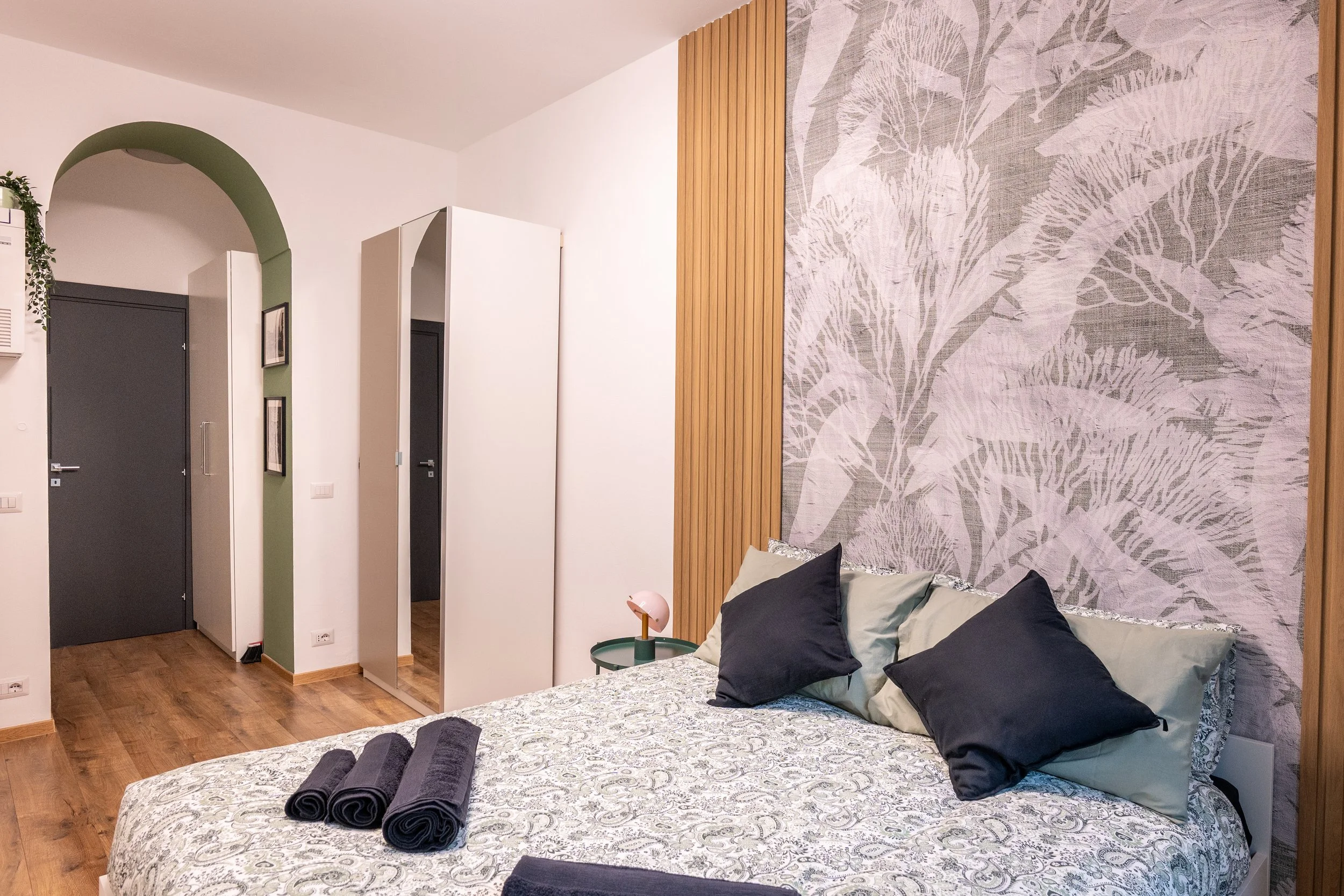 Camera matrimoniale con letto, cuscini neri e drappi, parete decorativa con foglie, lampada rosa, armadio bianco, pavimento di legno, porta grigia, pareti bianche e arco verde.