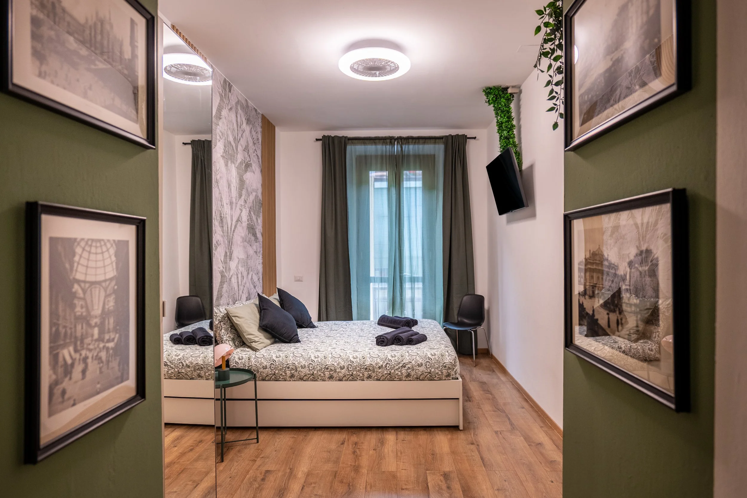 Camera da letto moderna con pareti bianche e decorazioni verdi, letto con cuscini e asciugamani, finestra con tende verdi, TV montato su parete, pavimento di legno.