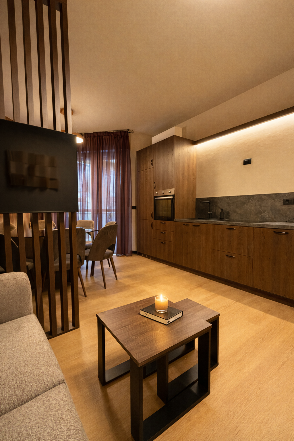 Interno di un appartamento con cucina a vista, tavolo con sedie, divano e una pianta, illuminazione calda e tende marroni.