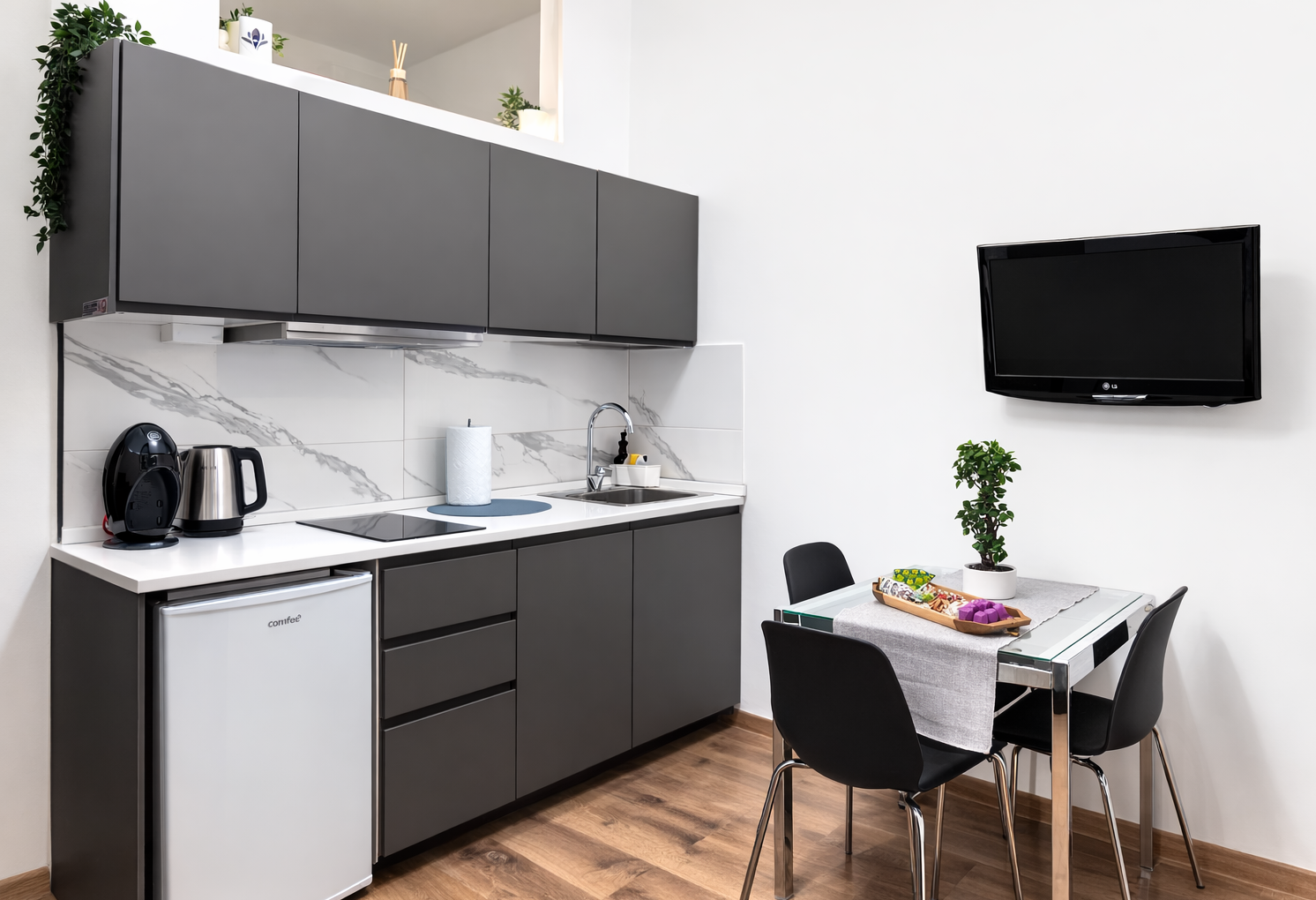 Cucina moderna con mobili grigi, piano di lavoro bianco, piccoli elettrodomestici, tavolo con sedie nere, televisore a parete, piante e decorazioni.