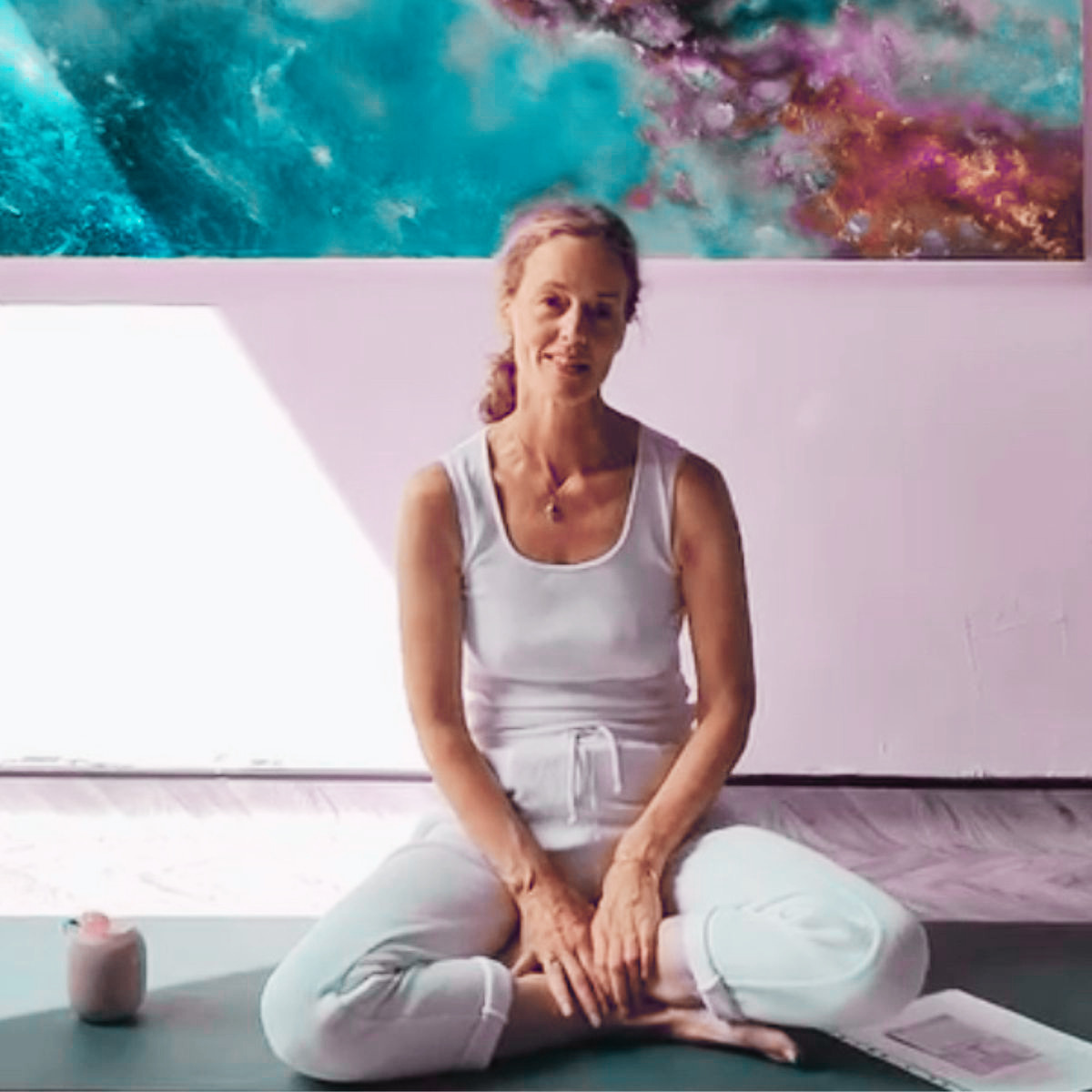 A middle-aged woman practicing breathwork in yoga studio, sitting cross-legged on a yoga mat with a small candle and a tablet nearby, in front of an abstract colourful painting.