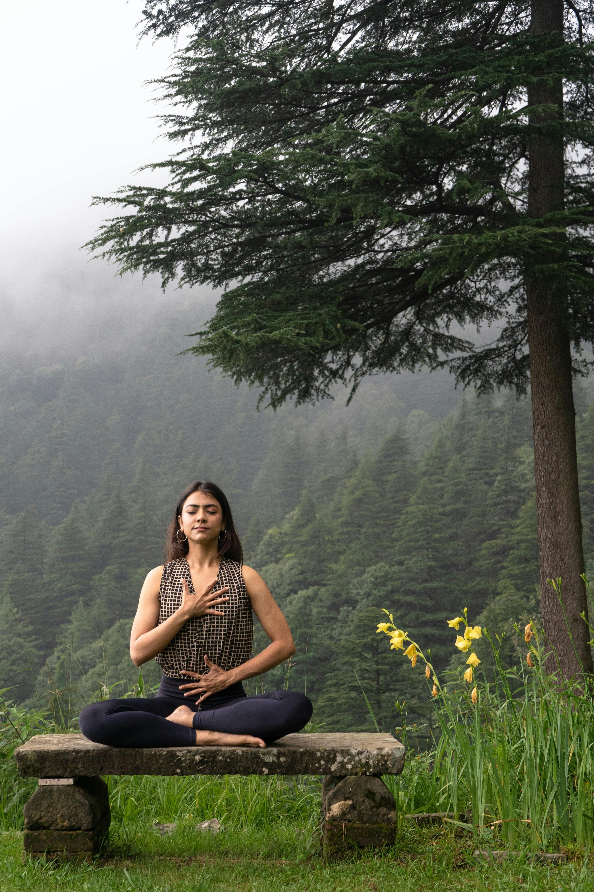 A woman practicing yoga outdoors on a stone platform in a lush green forest with fog, surrounded by yellow flowers.