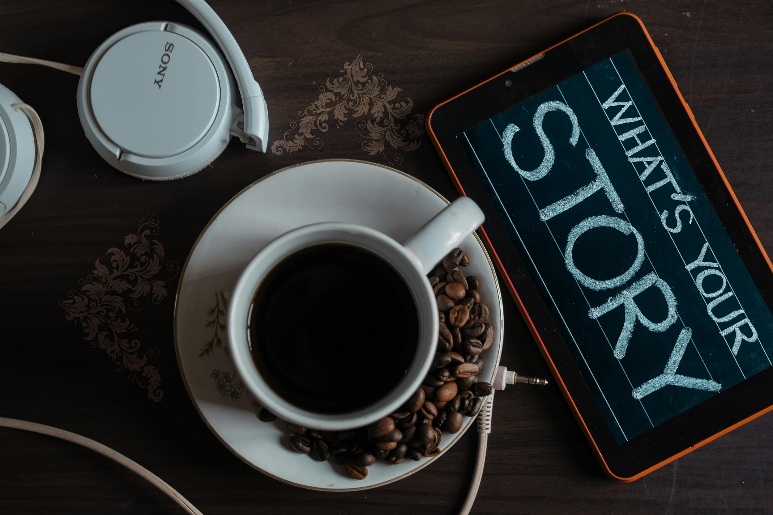 A top-down view of a wooden table with a cup of black coffee, a bowl of coffee beans, a pair of white Sony headphones, and a smartphone displaying a chalkboard-style sign that reads "What's Your Story".