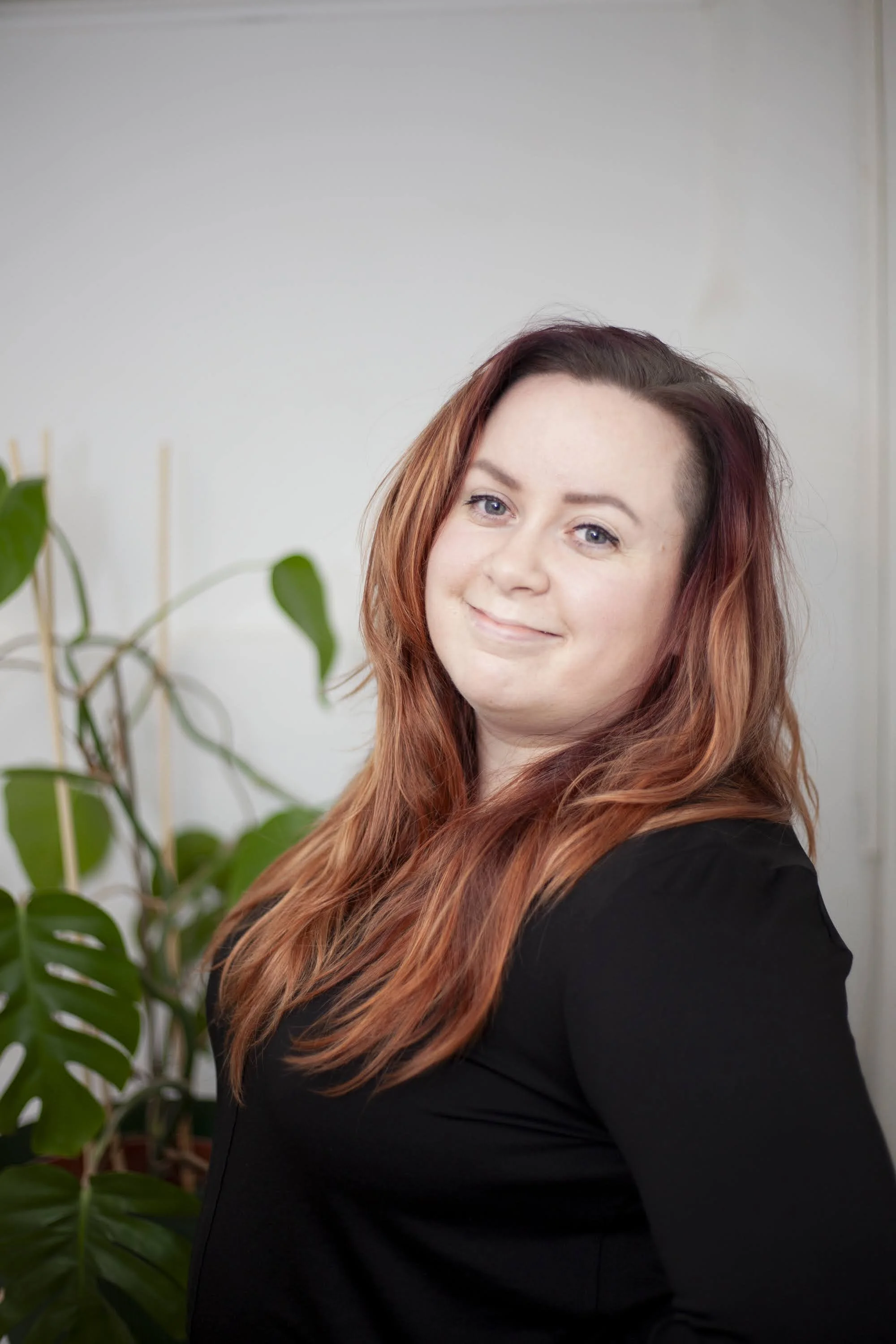 Kirjanpitäjä, joka hymyilee Lotteri Oy:n brändikuvauksissa. Hän on vaaleaihoinen nainen, ruskea punertava hiustenväri, musta paita, hymyilee kameralle, vihreitä lehtiä taustalla.