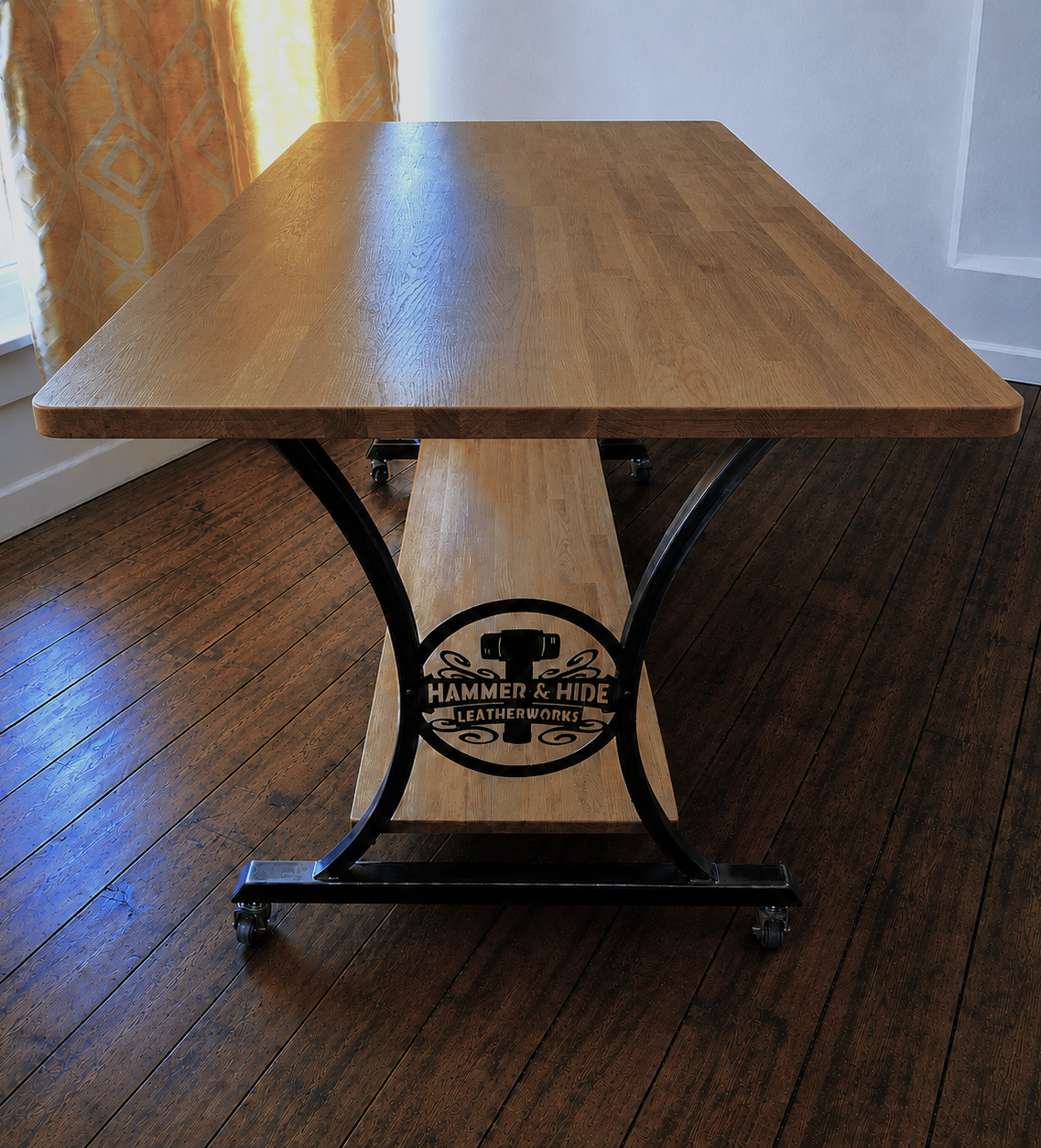 A custom made steel legged table with solid oak worktop, featuring a logo that reads 'Hammer & Hide Leatherworks.' The table is positioned on a dark wooden floor next to a window with yellow and gold patterned curtains.