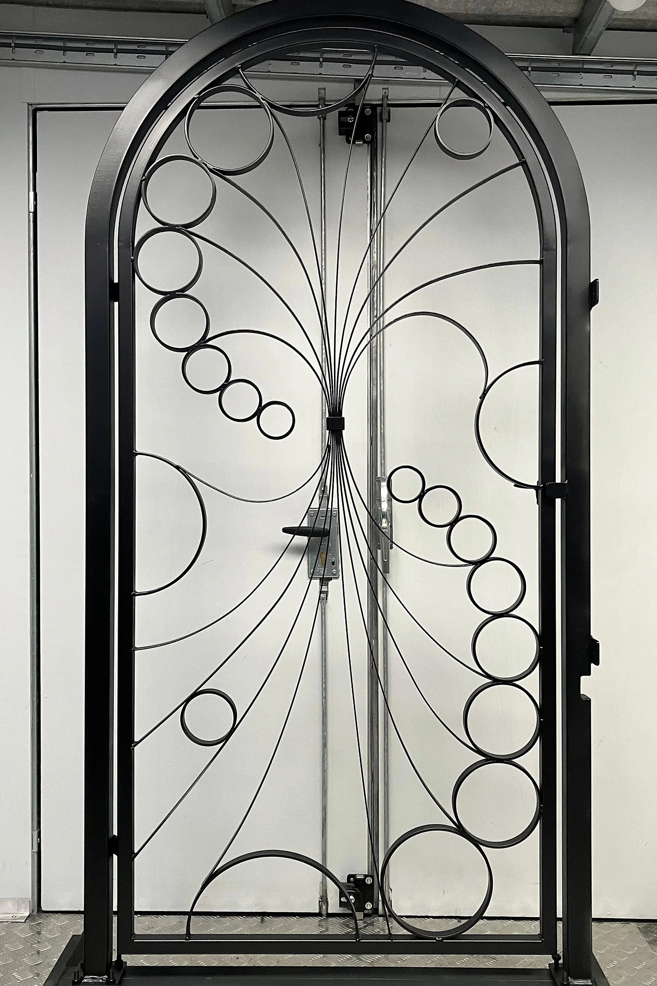 Metal decorative gate with circular patterns, mounted on a black frame, standing in front of a plain white wall.
